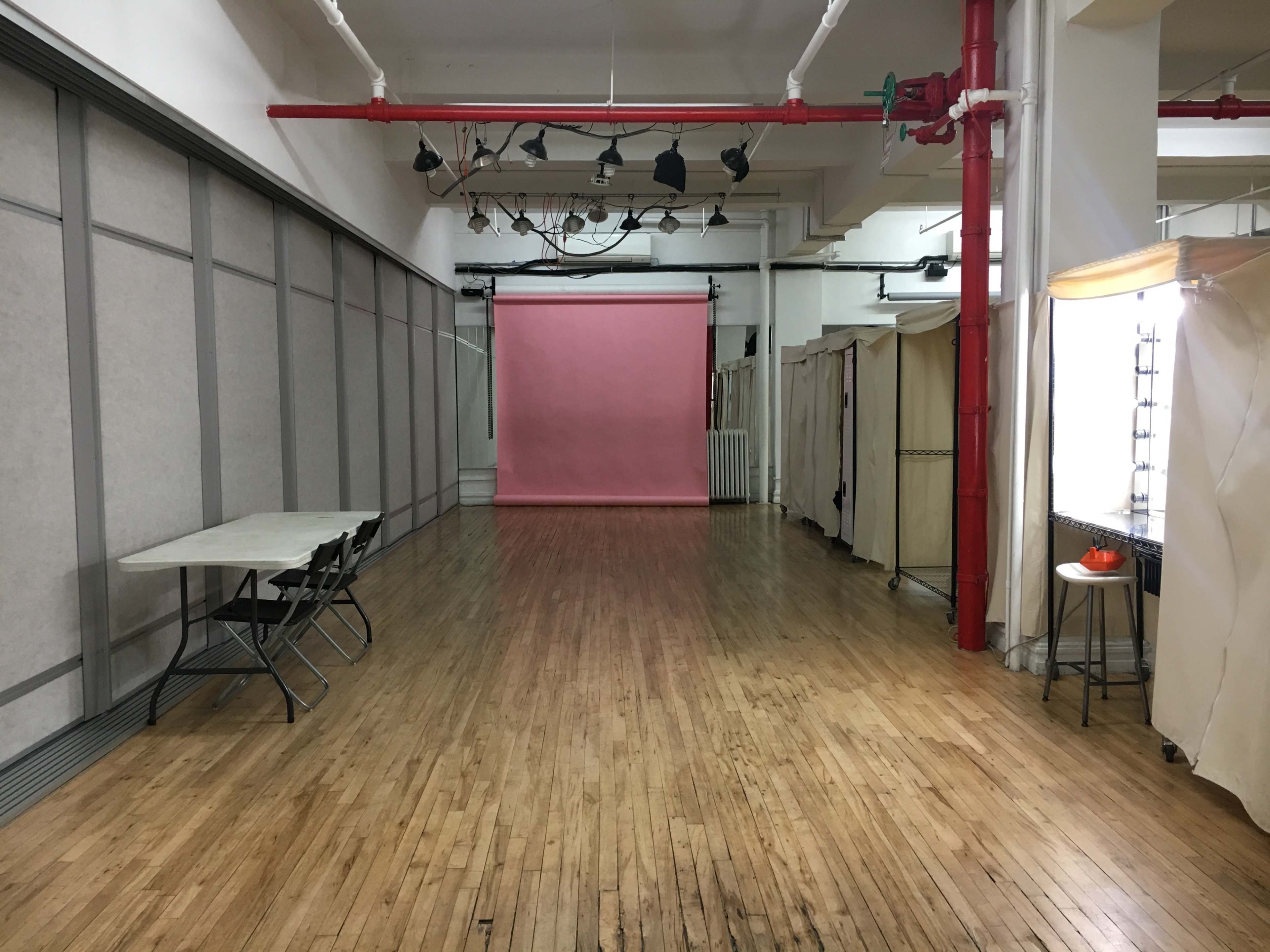 The image shows a spacious, empty studio with wooden floors, a pink backdrop at one end, and several partitioned areas along the sides.