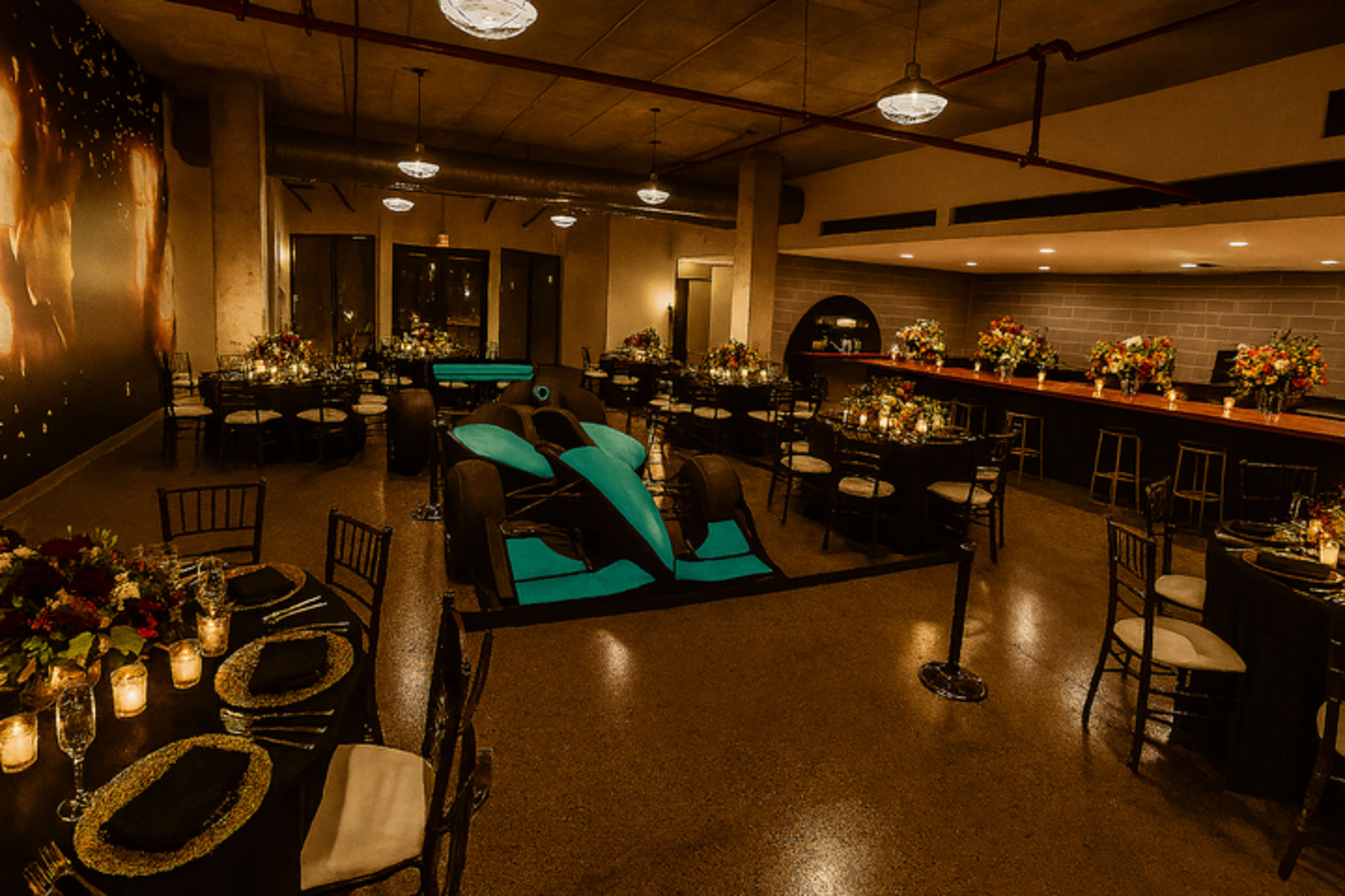 The image shows an elegant event space with round tables adorned with floral centerpieces and a sleek blue racing car prominently displayed in the center.