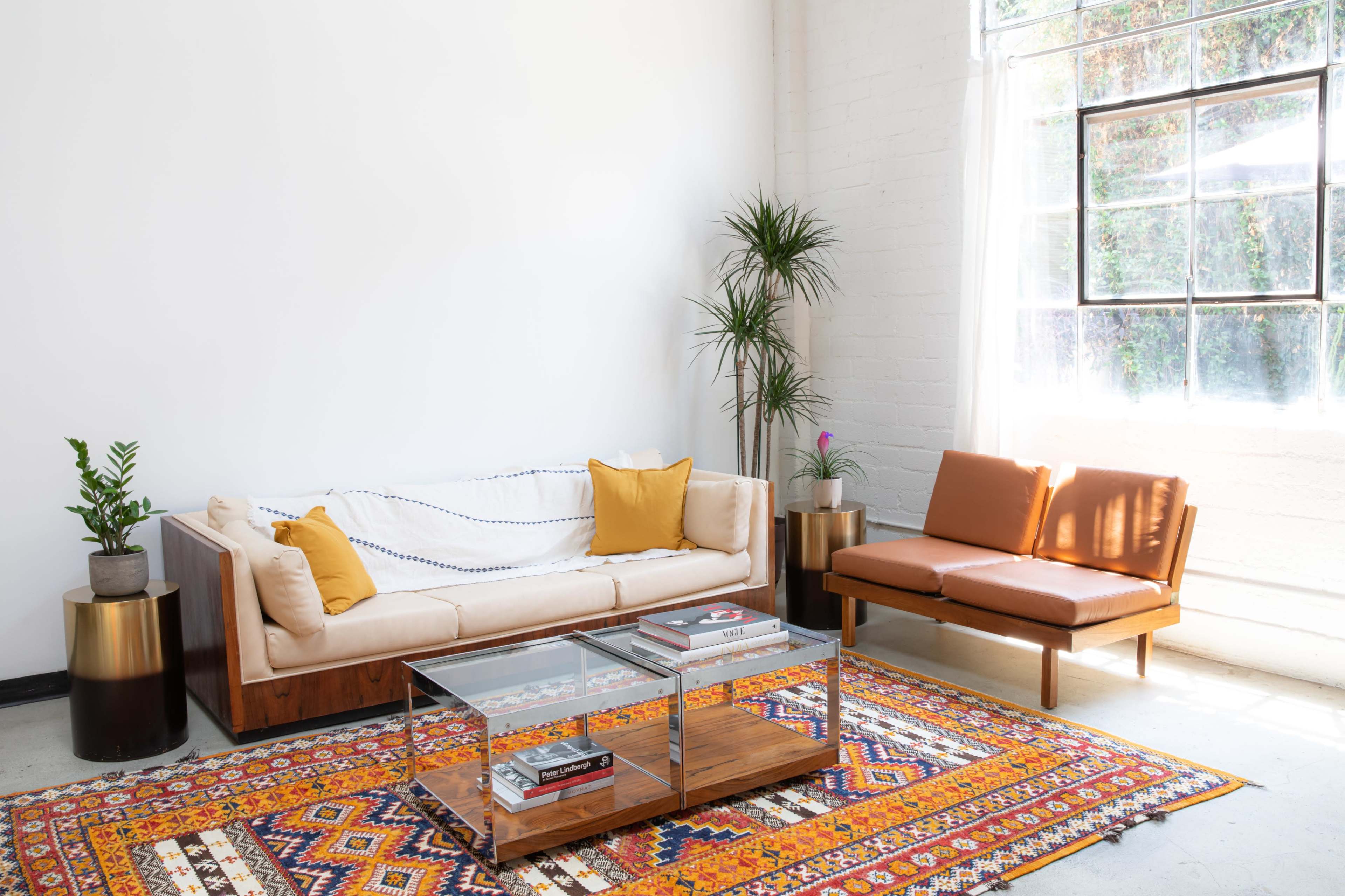A living room features a beige sofa with yellow cushions, a brown leather chair, a glass coffee table, and a colorful patterned rug, all against a backdrop of large windows and potted plants.