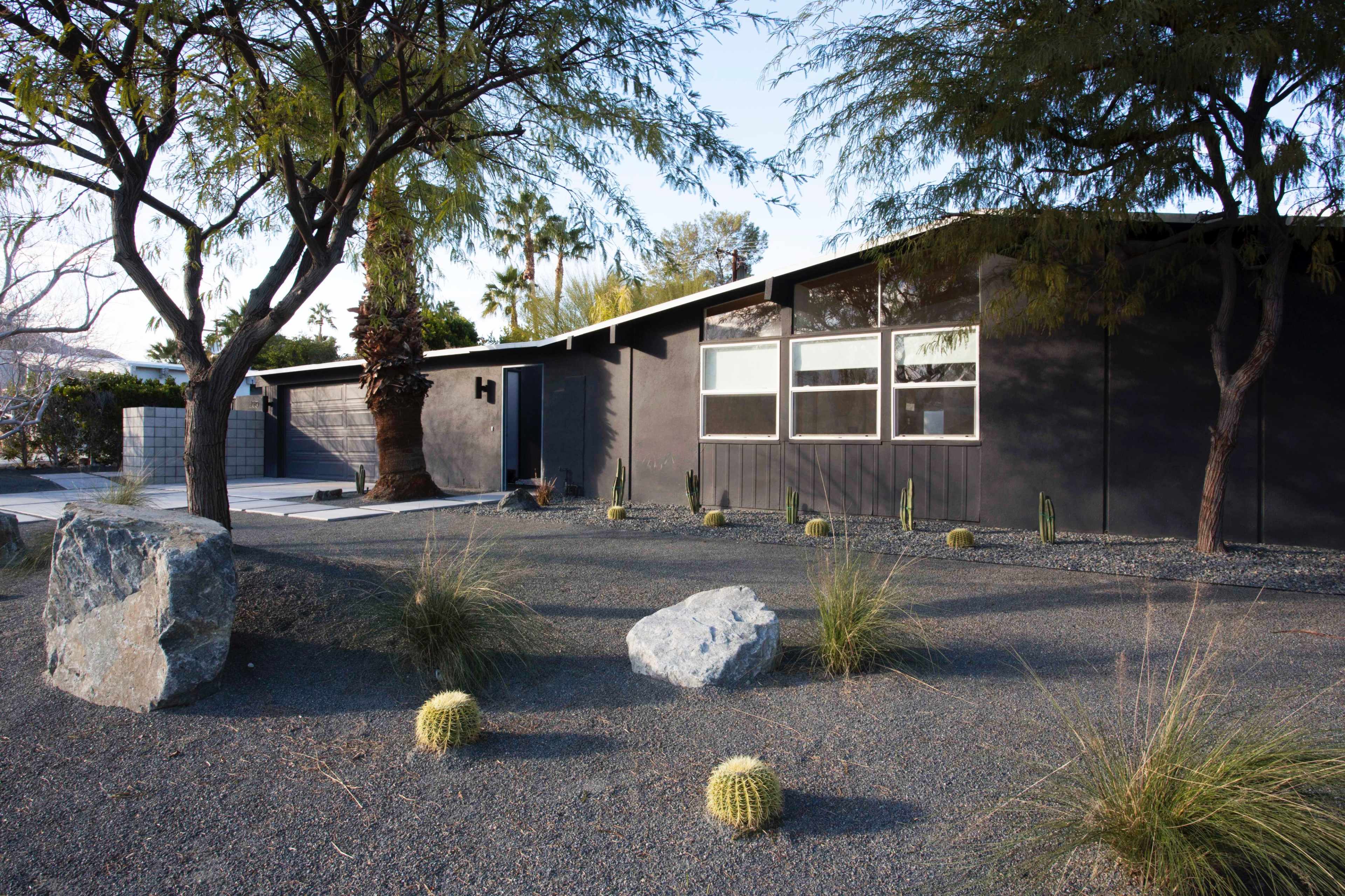 A modern house with large windows is surrounded by gravel landscaping, cacti, and various trees under clear skies.
