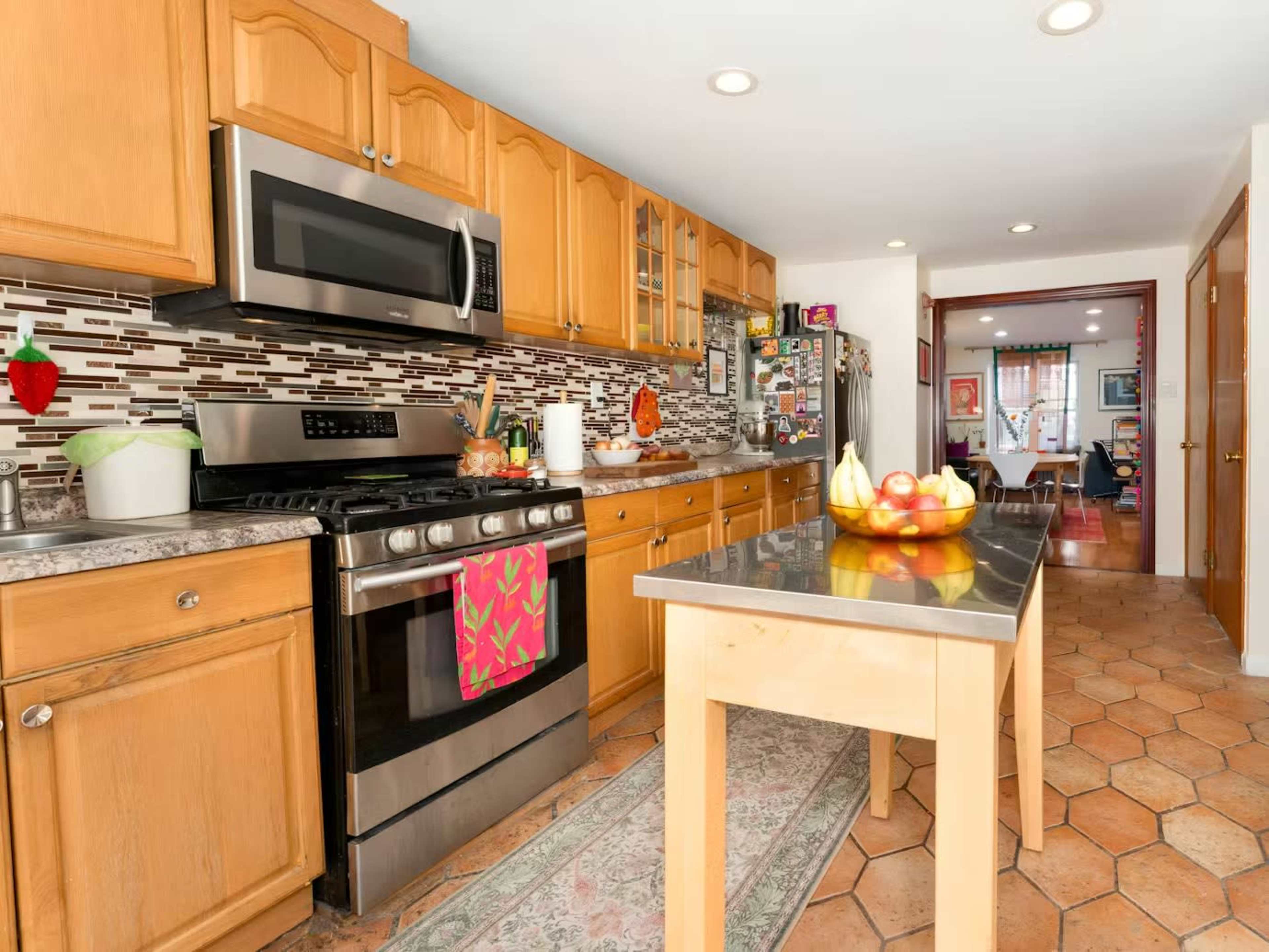 The image shows a modern kitchen with wooden cabinets, stainless steel appliances, and a central island featuring a bowl of fruits.