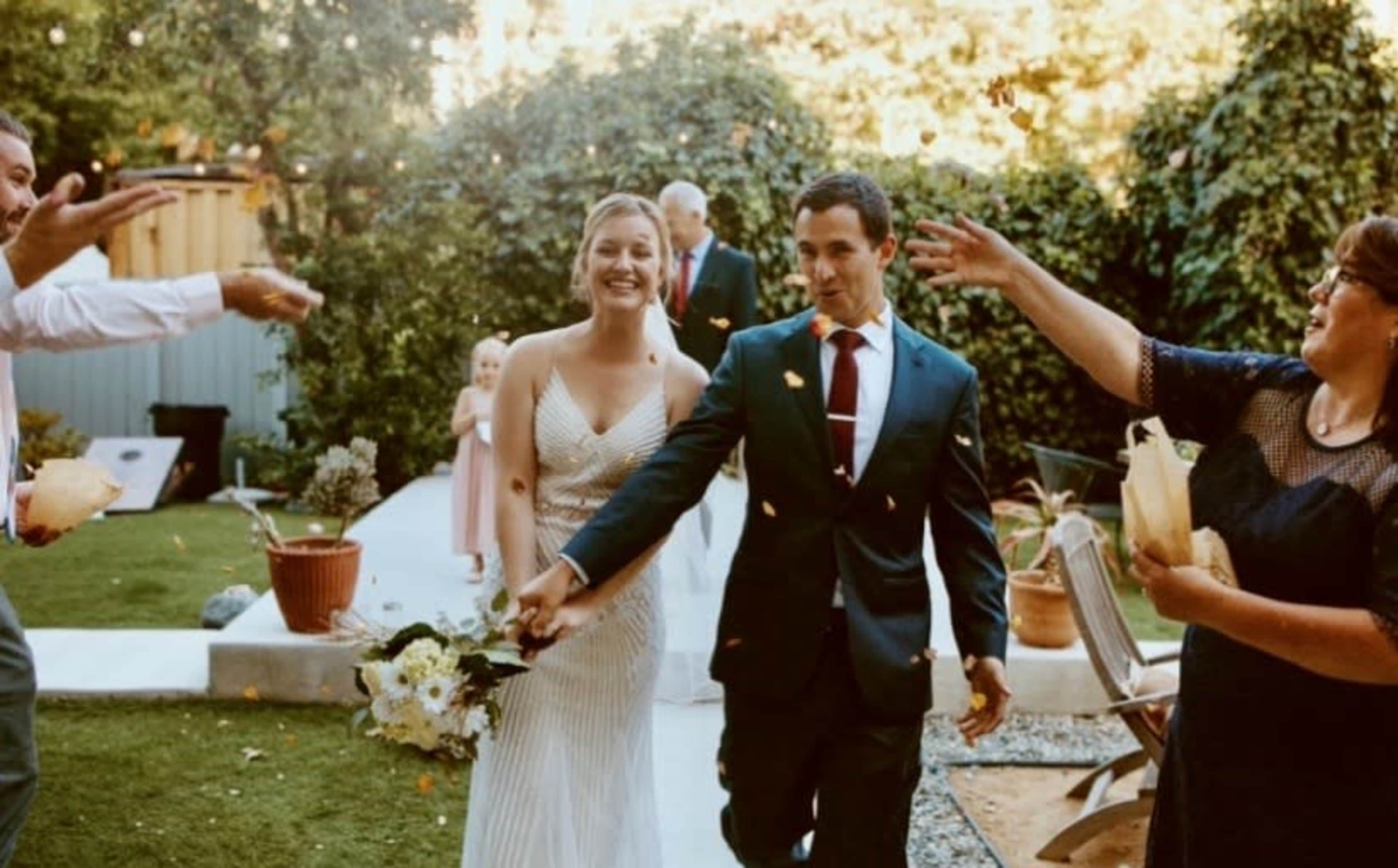 A couple walks hand in hand down a path as guests throw petals in celebration at an outdoor wedding.