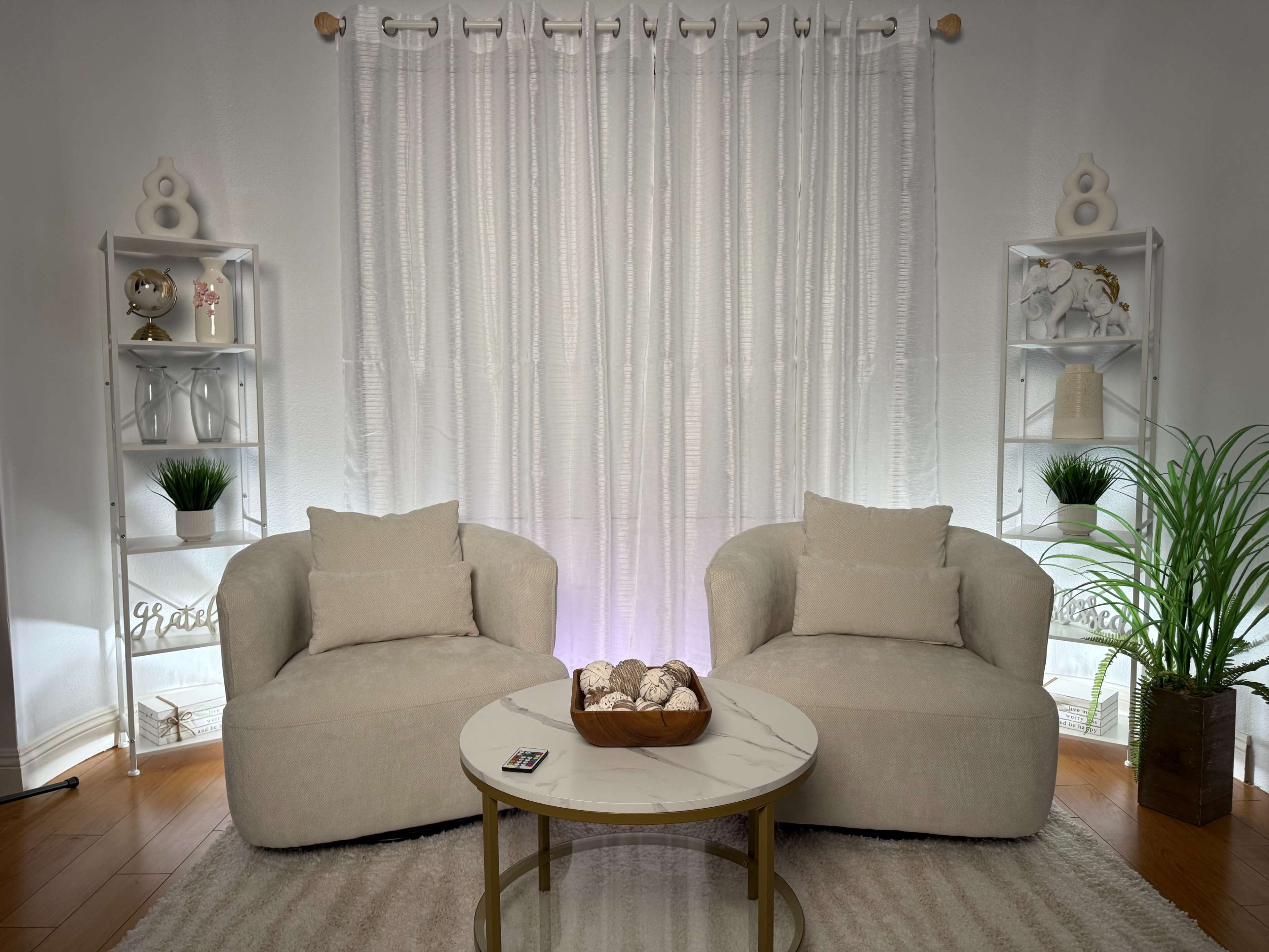 The image shows a cozy living room setting featuring two light-colored armchairs, a round coffee table with decorative items, and shelves with plants and ornaments against a backdrop of sheer white curtains.