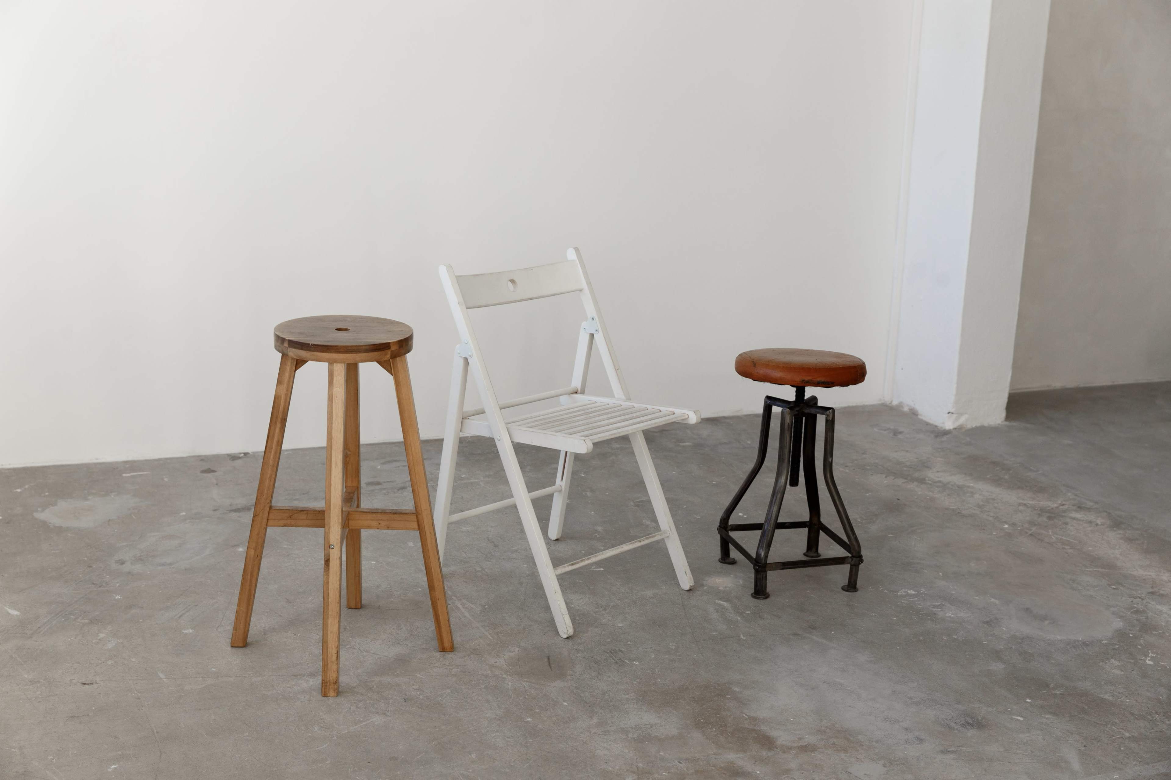 Three different stools: one wooden, one white folding chair, and one black metal stool, all placed on a gray concrete floor against a plain wall.