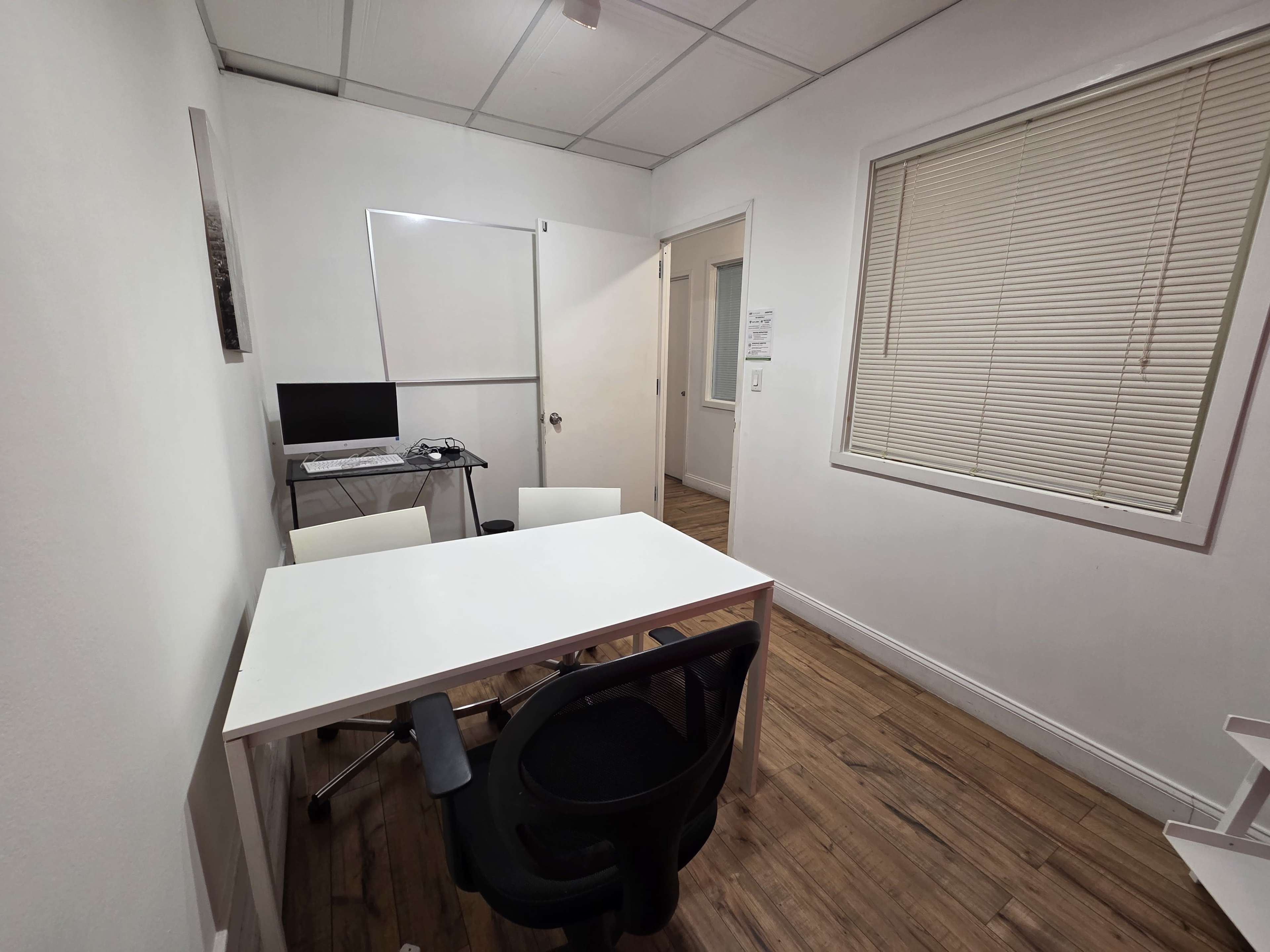 The image shows a small office space with a white table, three chairs, a computer on a desk, and a door leading to another room.