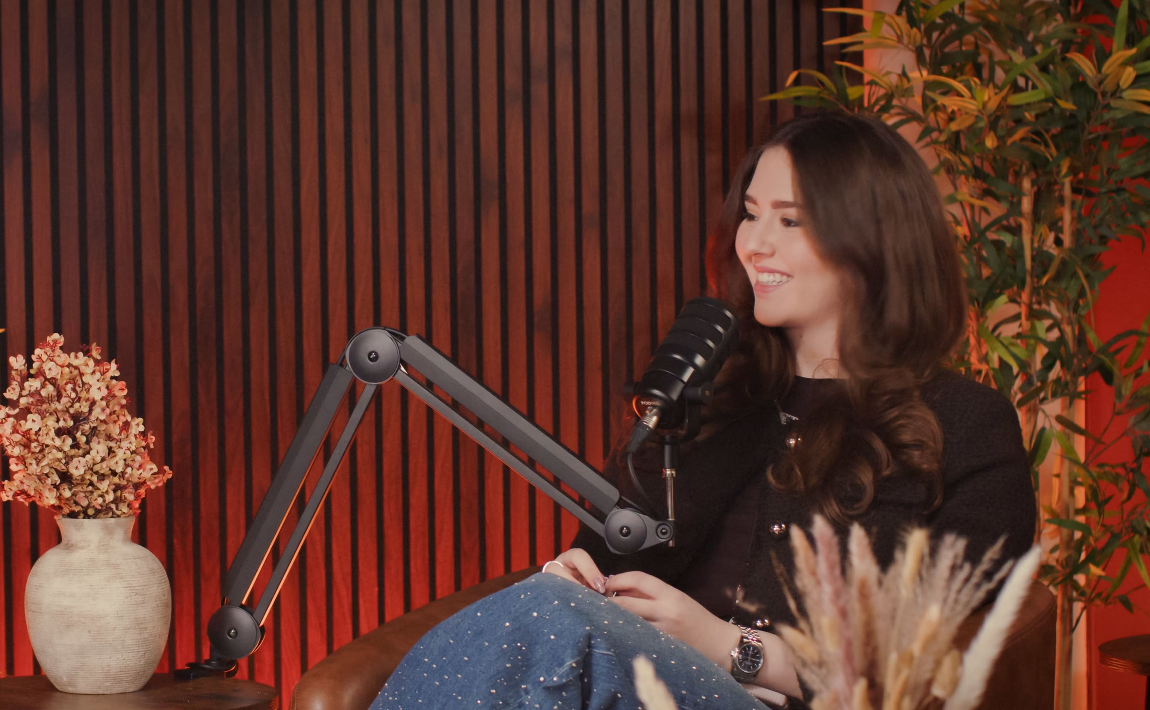 A person is seated in a cozy studio with a microphone on a stand, surrounded by decorative plants and a vase of dried flowers.
