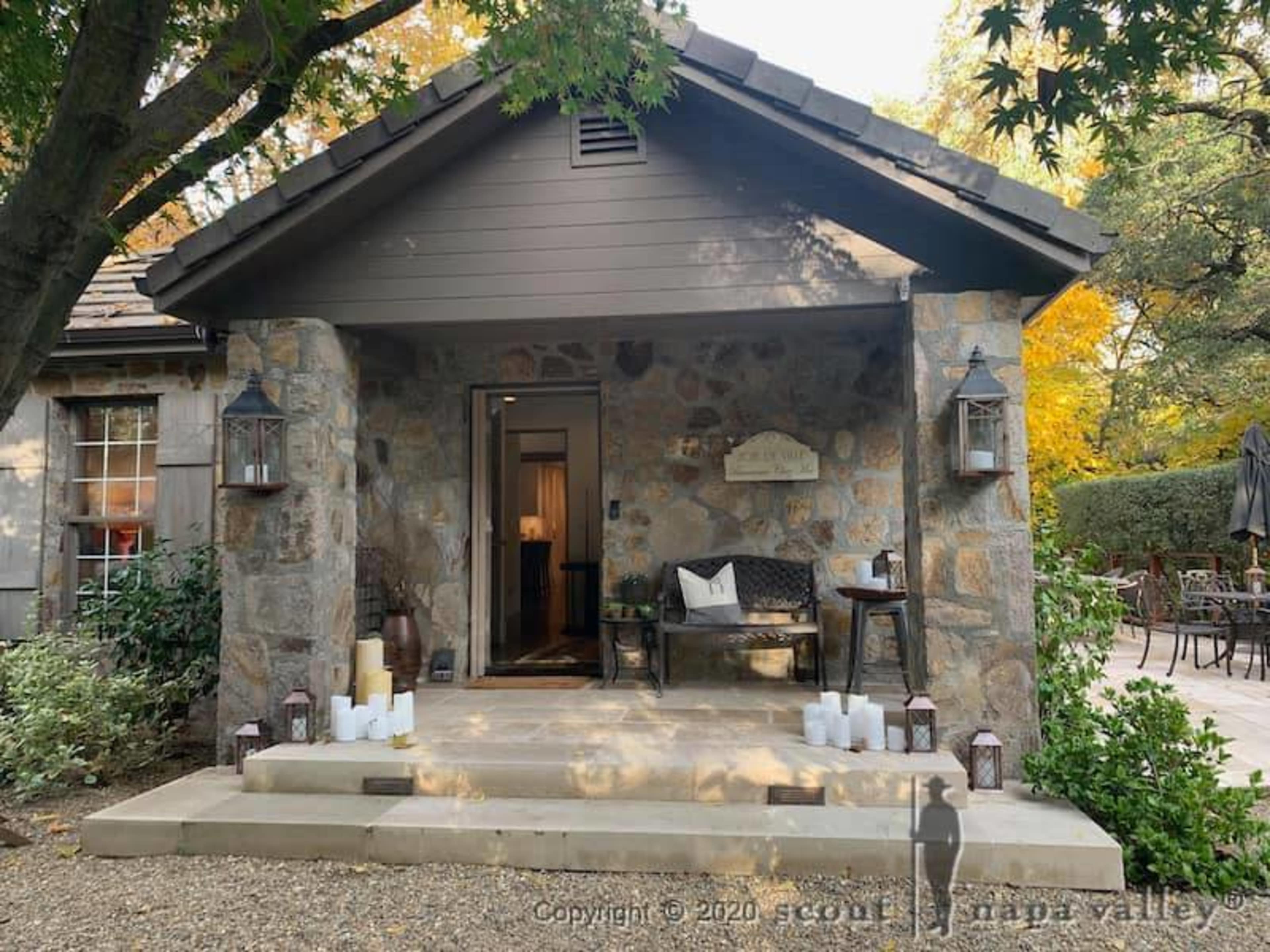 A stone cottage with a wooden porch, flanked by lanterns and white candles, is set amidst a landscaped garden.