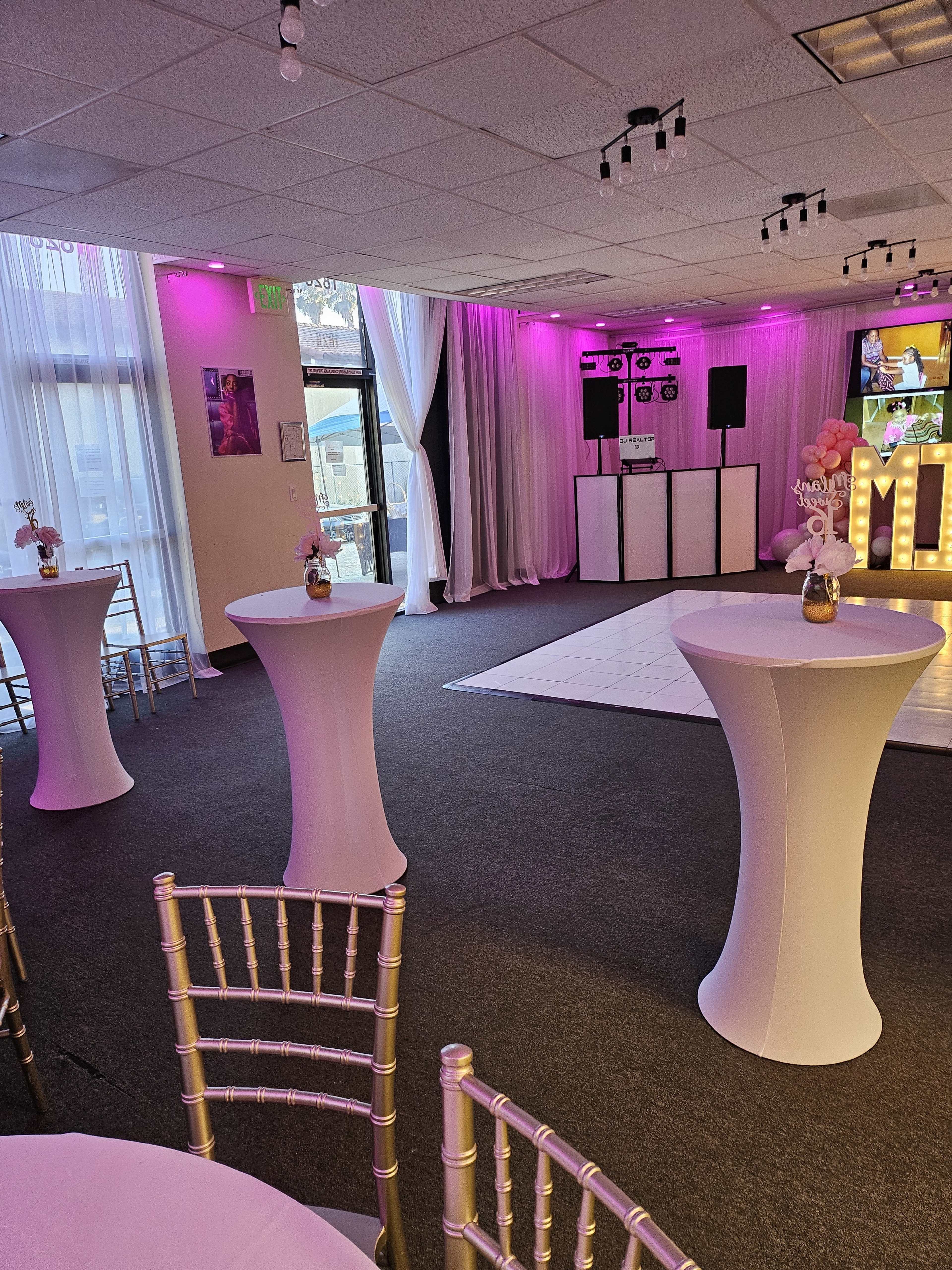 The image shows a spacious event venue with white, round tables surrounded by gold chiavari chairs, illuminated by pink lighting and featuring a dance floor.