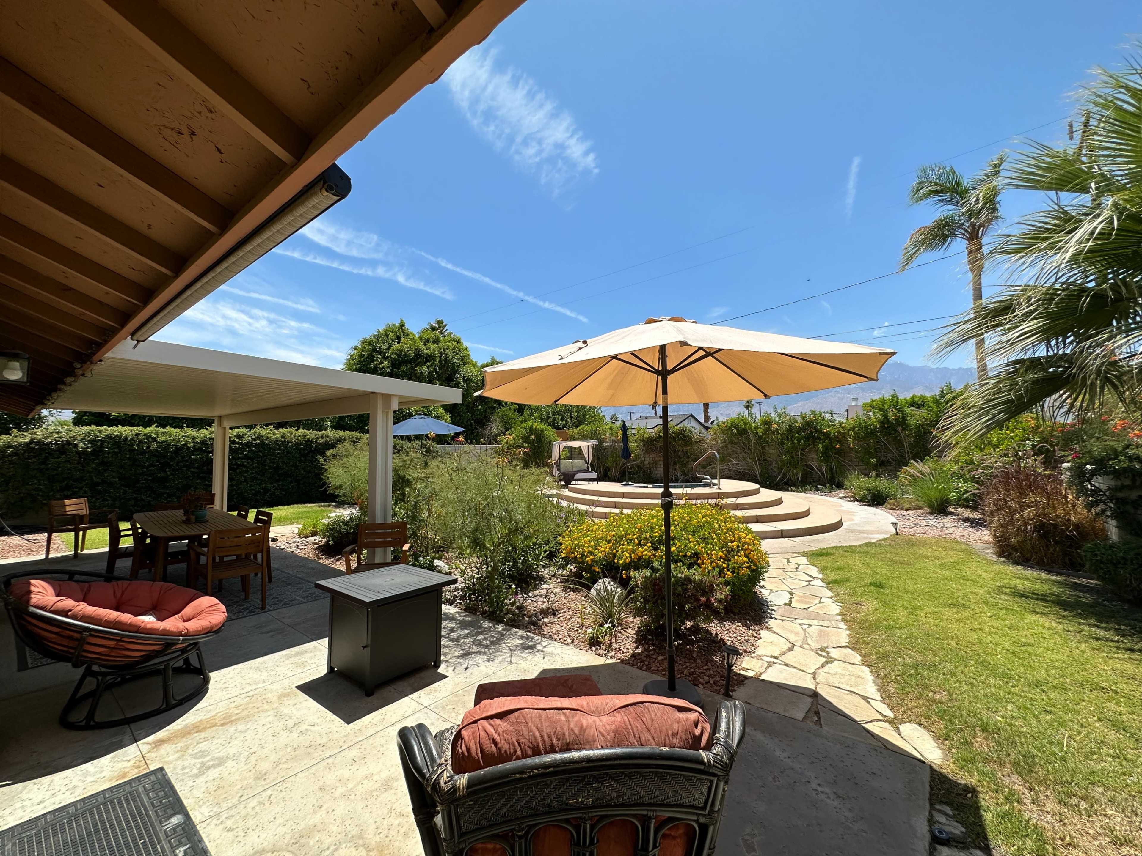 The image shows a landscaped backyard featuring a seating area with an umbrella, a stone pathway, and steps leading to a raised circular platform surrounded by greenery.