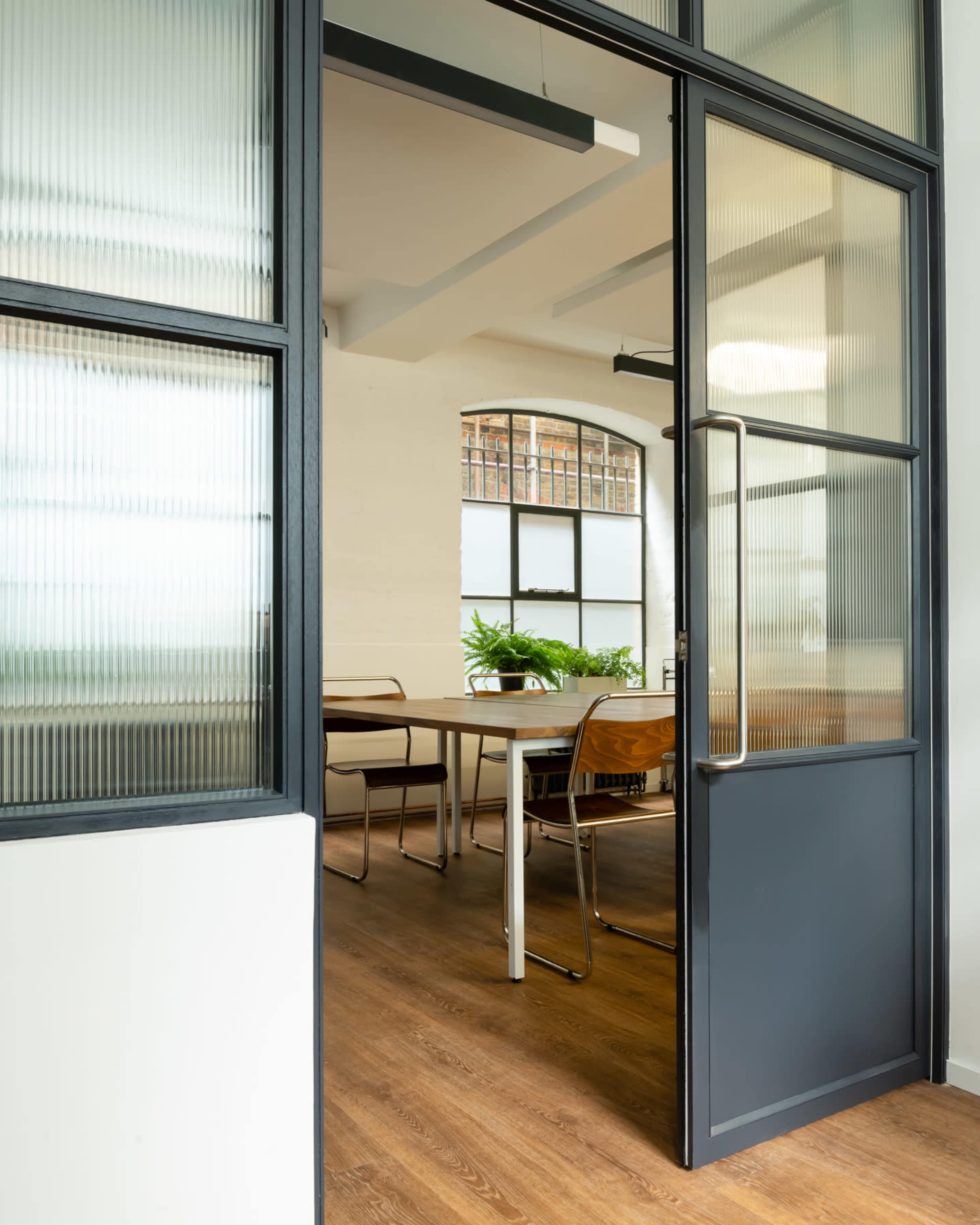 A modern office space with a wooden floor, featuring a glass door that opens to a meeting room with a table and chairs surrounded by plants.