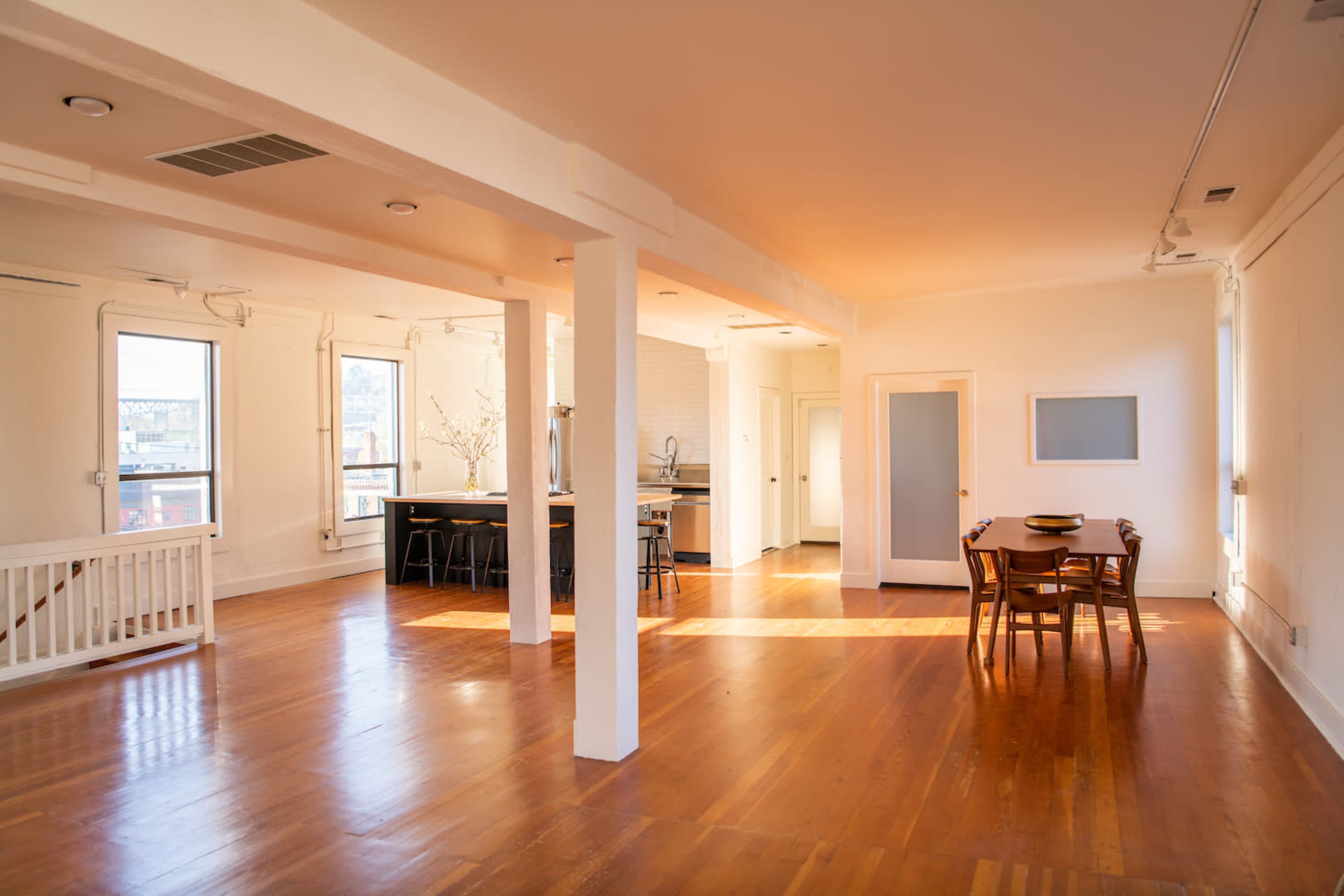 A spacious room with wooden floors, tall windows, and a dining area featuring a table with chairs.