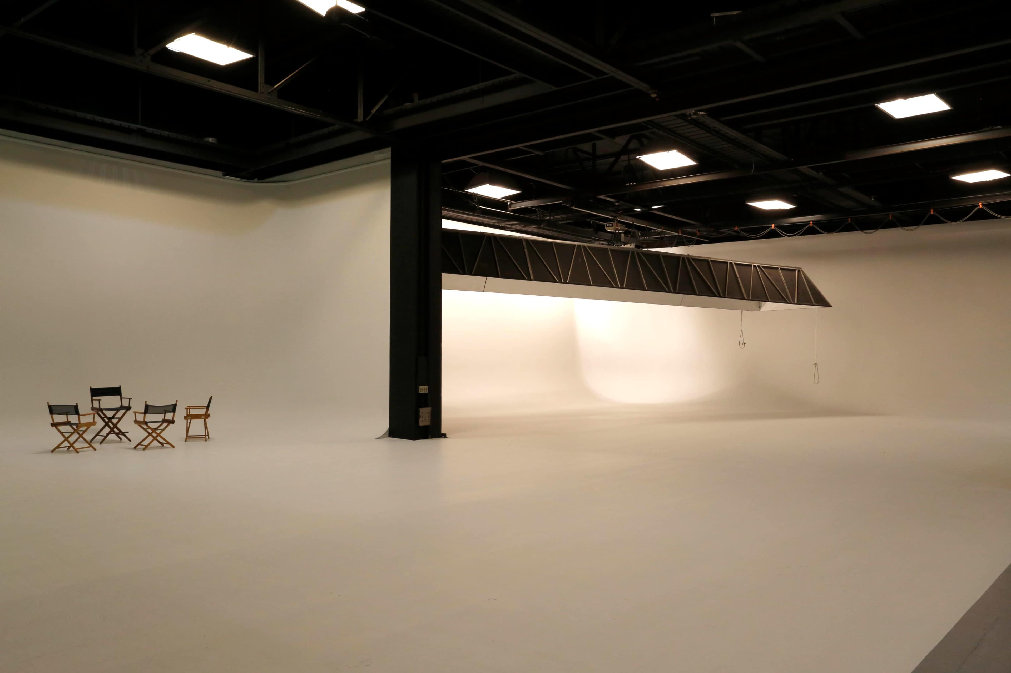 An empty film studio with a white backdrop, large space, and a few director's chairs arranged on one side.