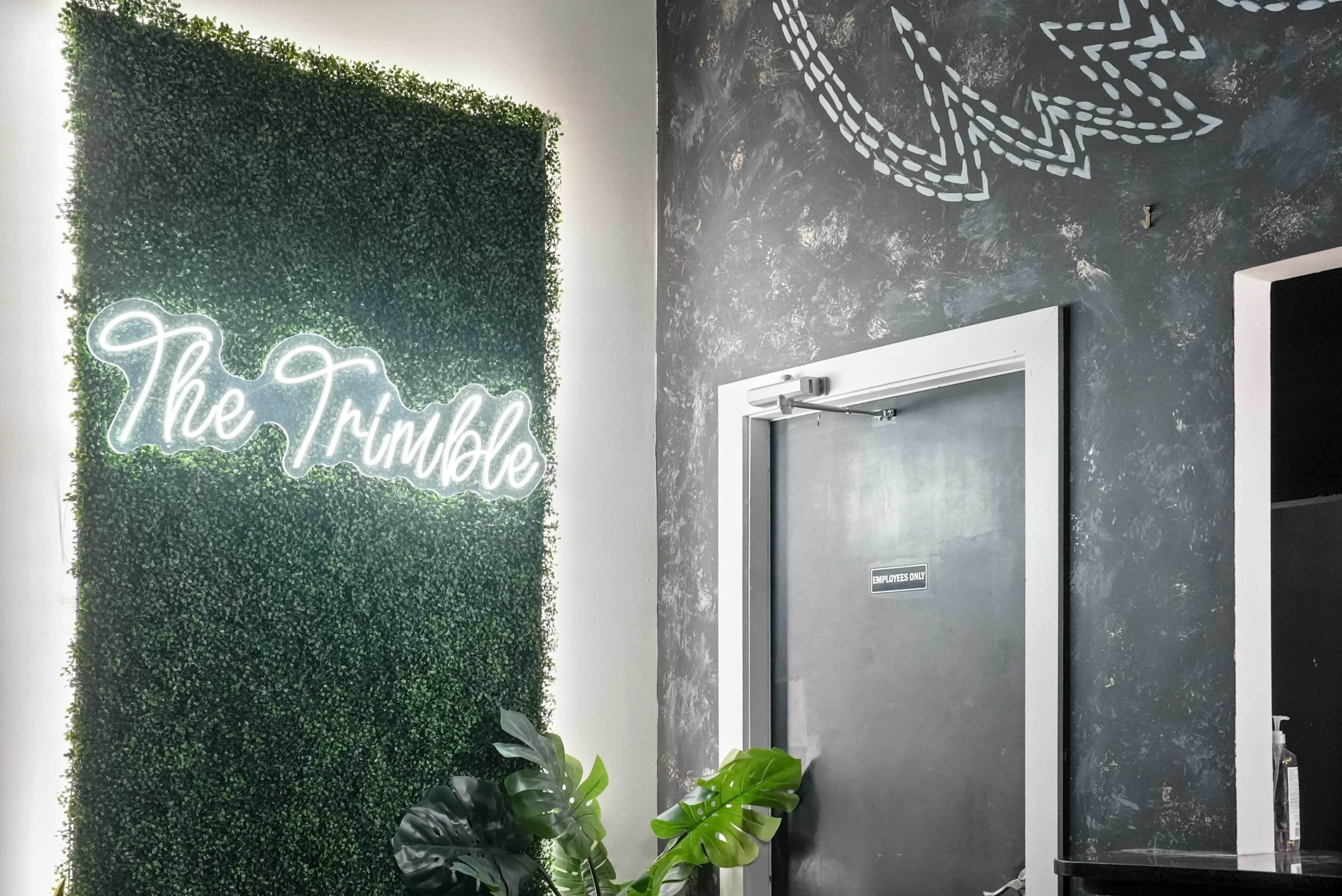 The image shows a wall covered in artificial greenery with the words "The Trimble" illuminated in neon, next to a door against a textured black wall.