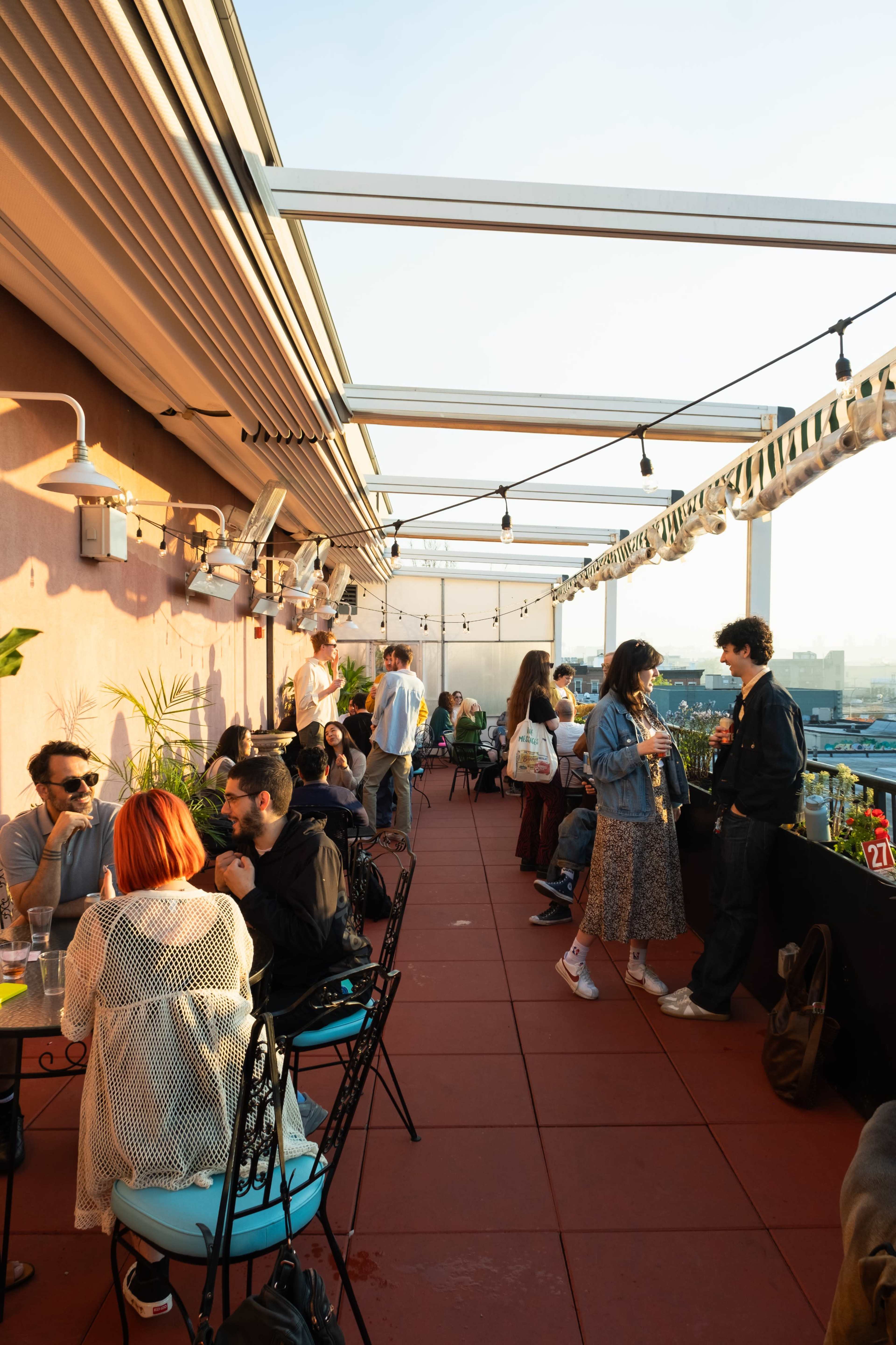 The image shows a vibrant rooftop bar scene with people socializing at tables and standing near a bar under string lights.