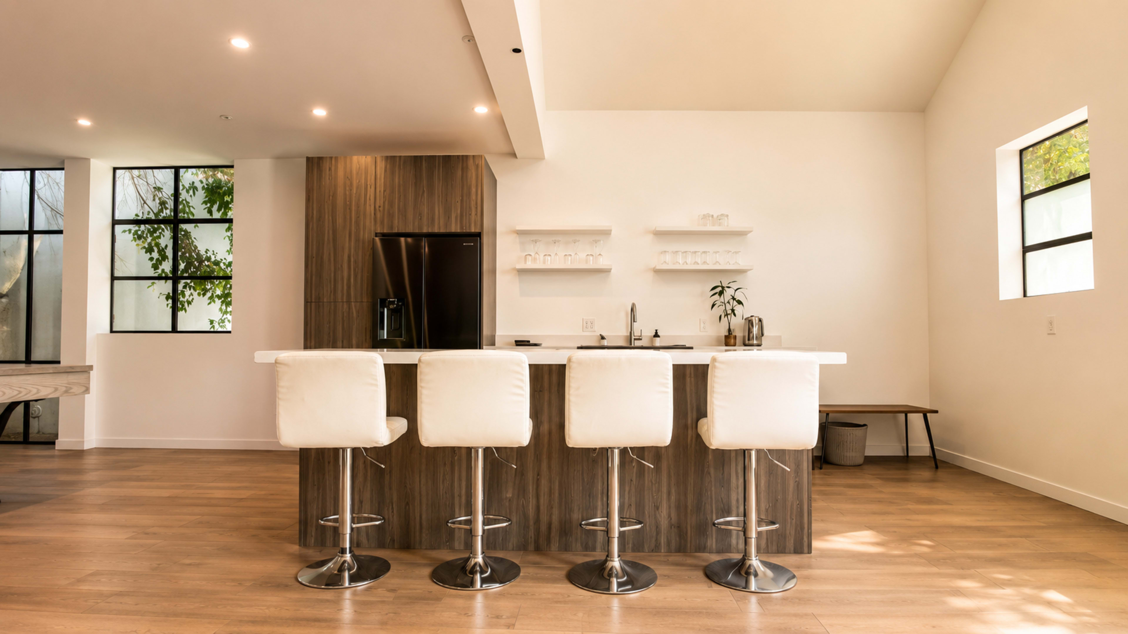 The image shows a modern kitchen with a central island featuring four bar stools, a dark refrigerator, and minimalistic decor.