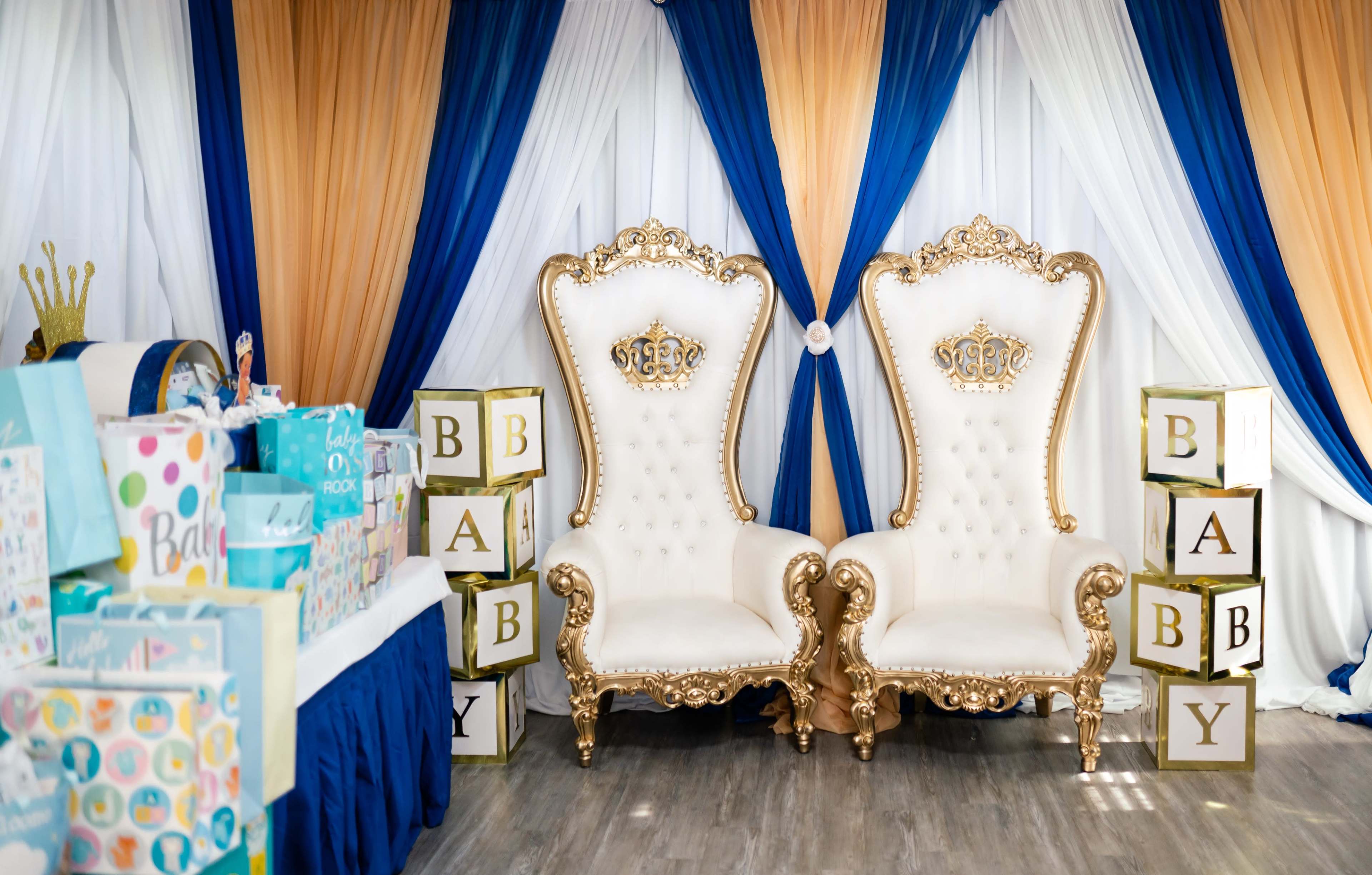 The image shows two ornate white and gold throne chairs positioned against a backdrop of blue and orange drapery, with a table of decorated gift boxes nearby.