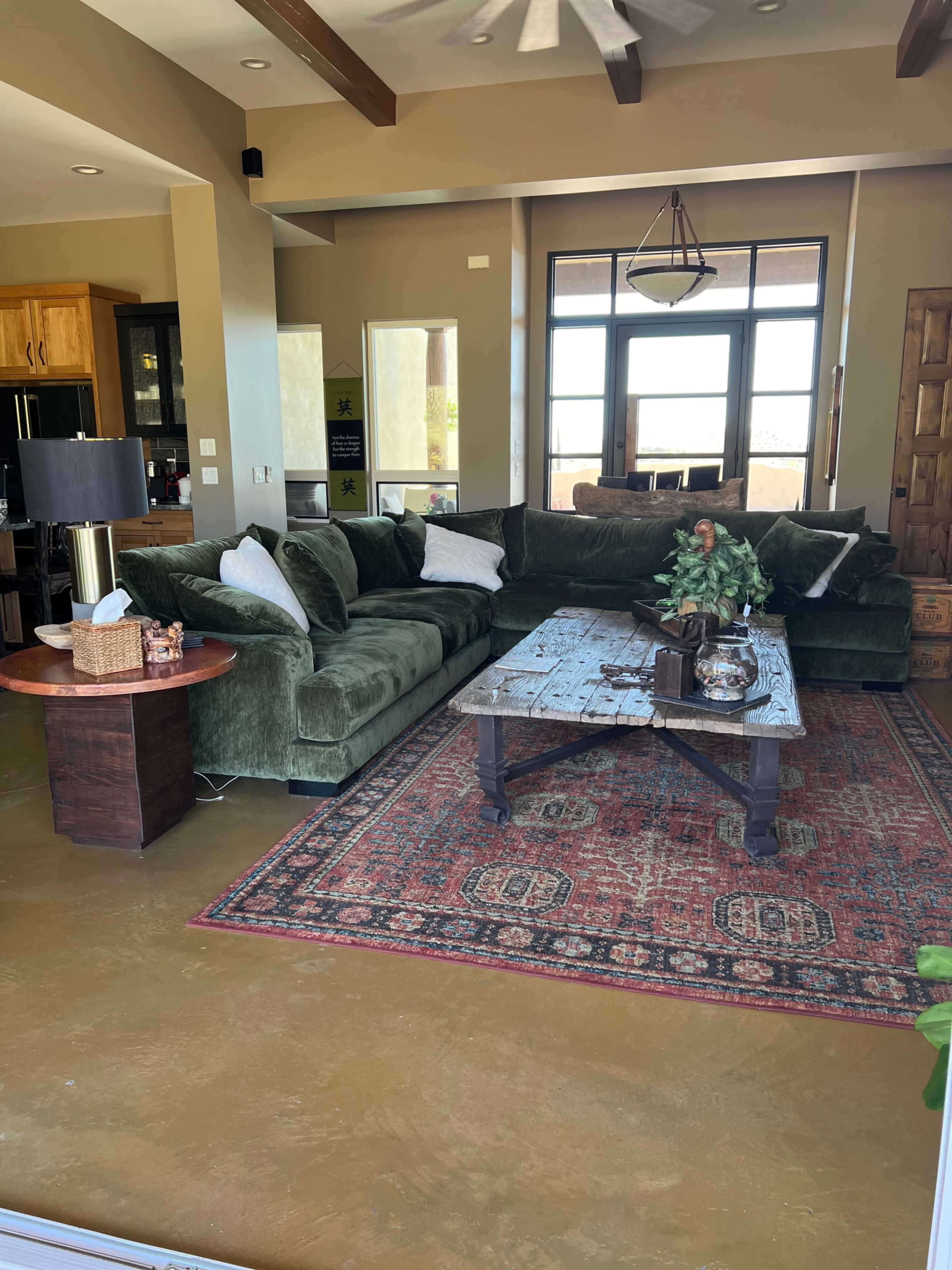 The living room features a large green sectional sofa, a wooden coffee table with decorative items, and a colorful area rug over a polished concrete floor.