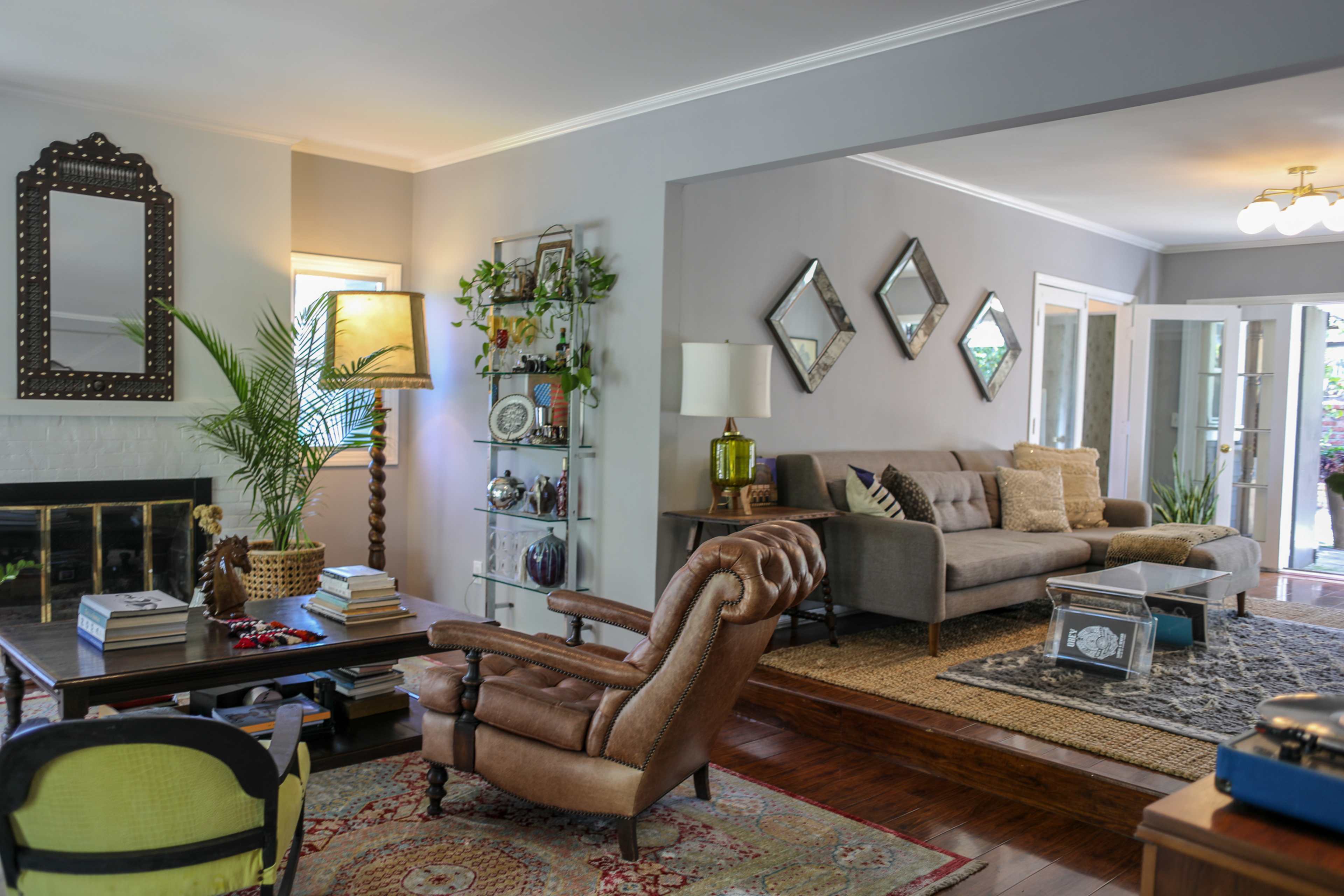 A cozy living room features a gray sofa, a wooden coffee table, a decorative shelf, and a vintage armchair, surrounded by plants and framed mirrors on the walls.