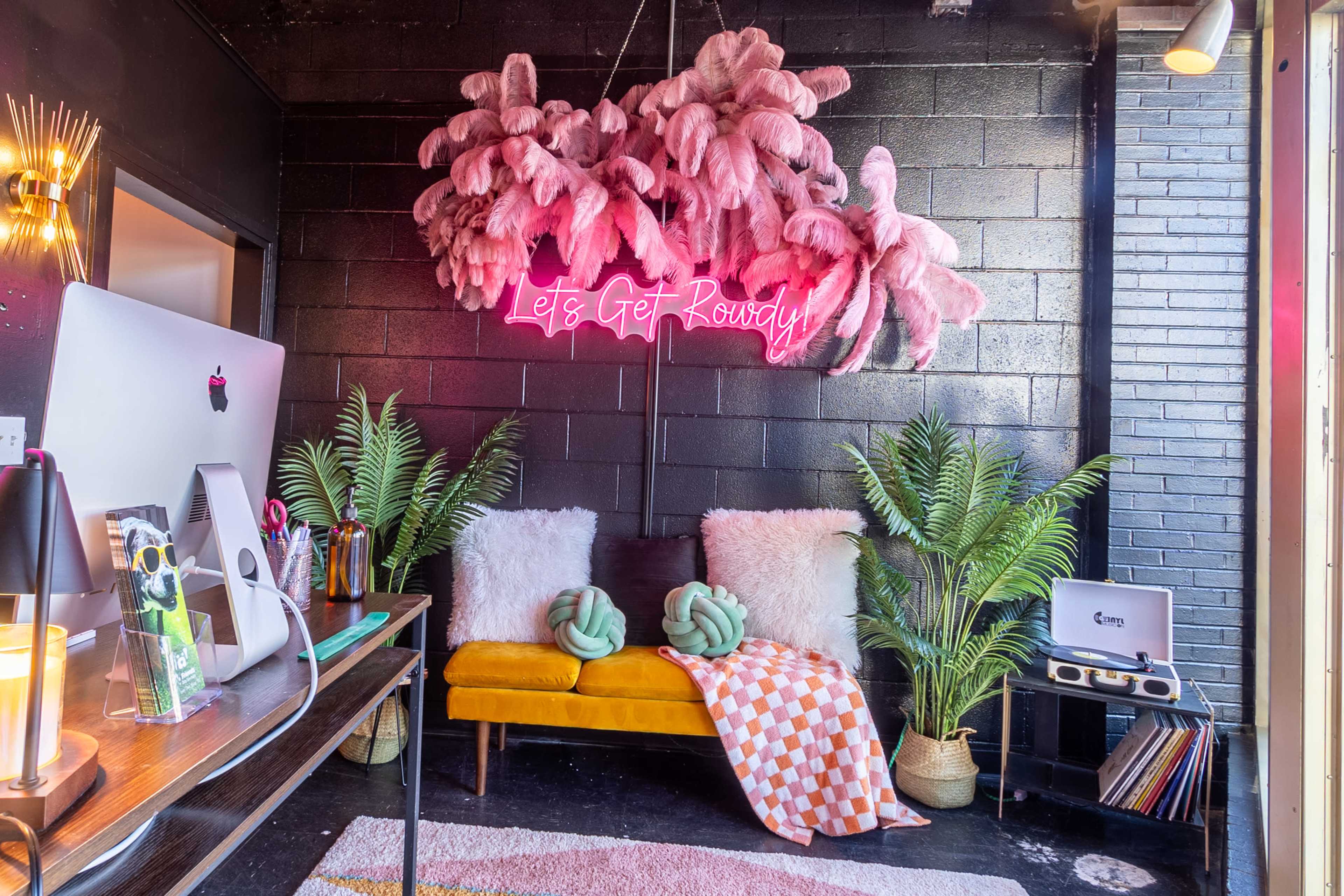 A cozy, stylish interior features a yellow couch with decorative pillows, a neon sign that says "Let's Get Rowdy," and plants arranged around a workspace with a computer and vinyl records.