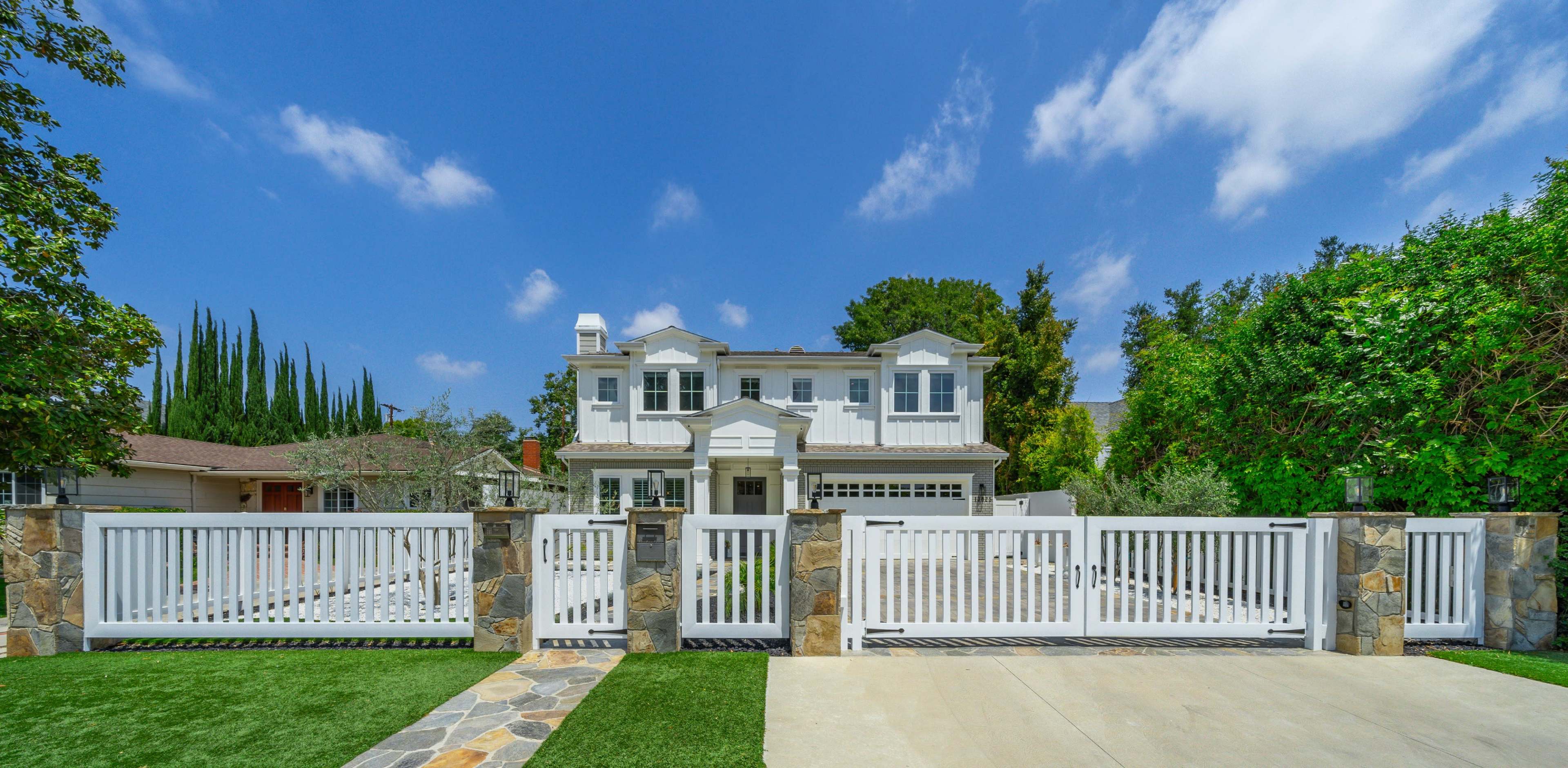 A two-story white house with a front porch and a landscaped yard is enclosed by a white fence and gate.