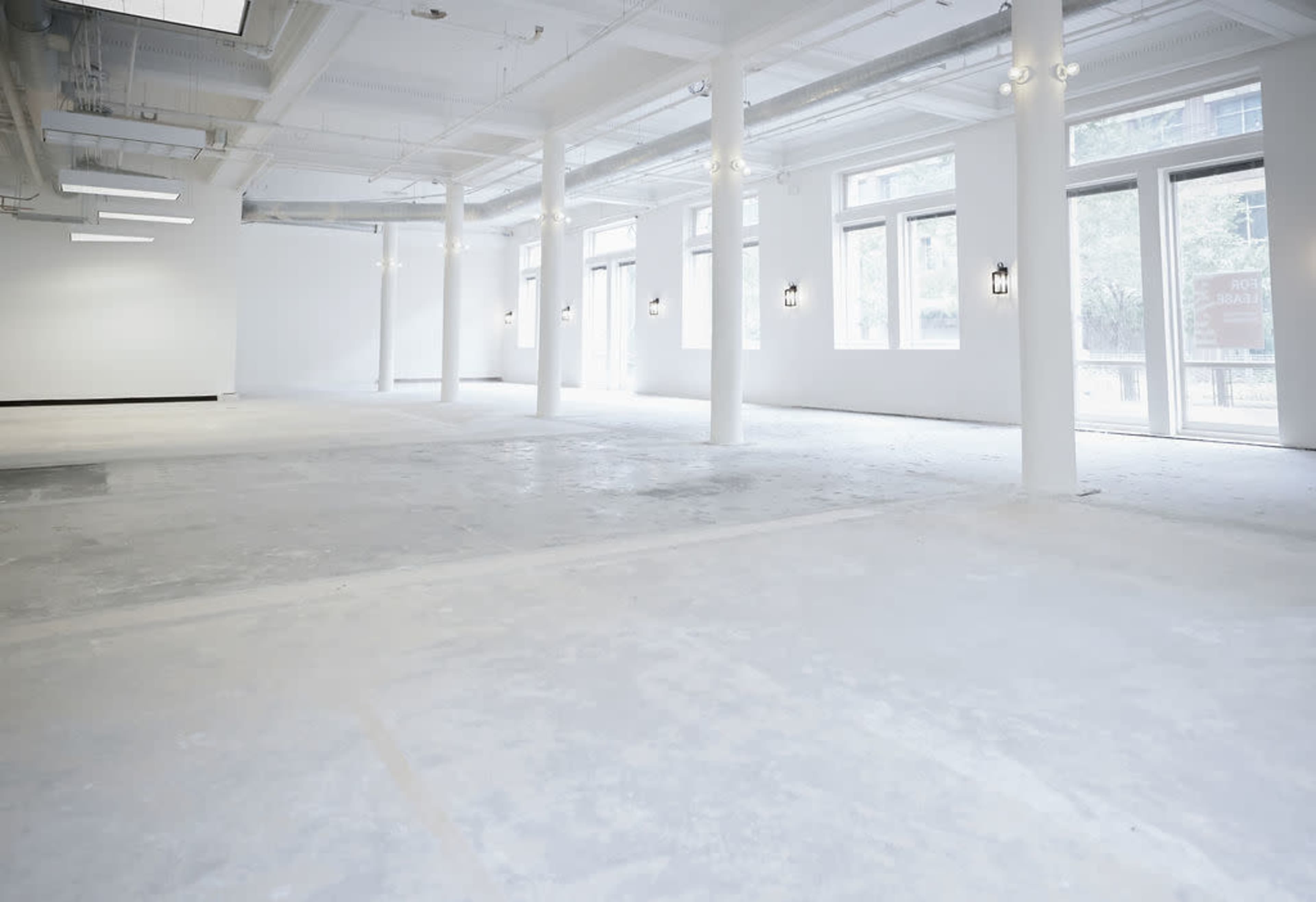 The image shows a large, empty room with a concrete floor, bright white walls, and several windows that let in natural light.
