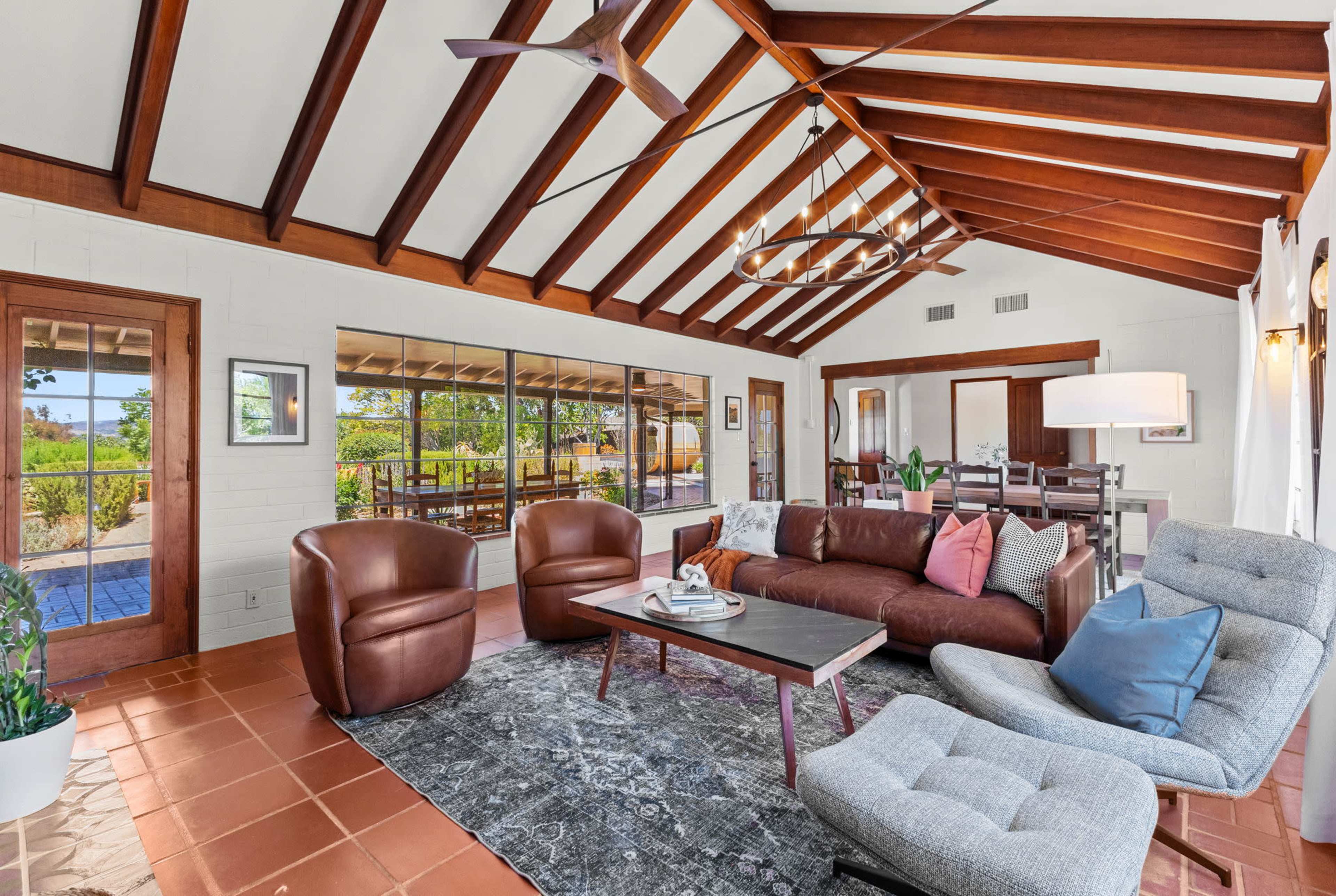 A bright living room features a brown leather sofa, two matching armchairs, a coffee table, and large windows overlooking a garden.