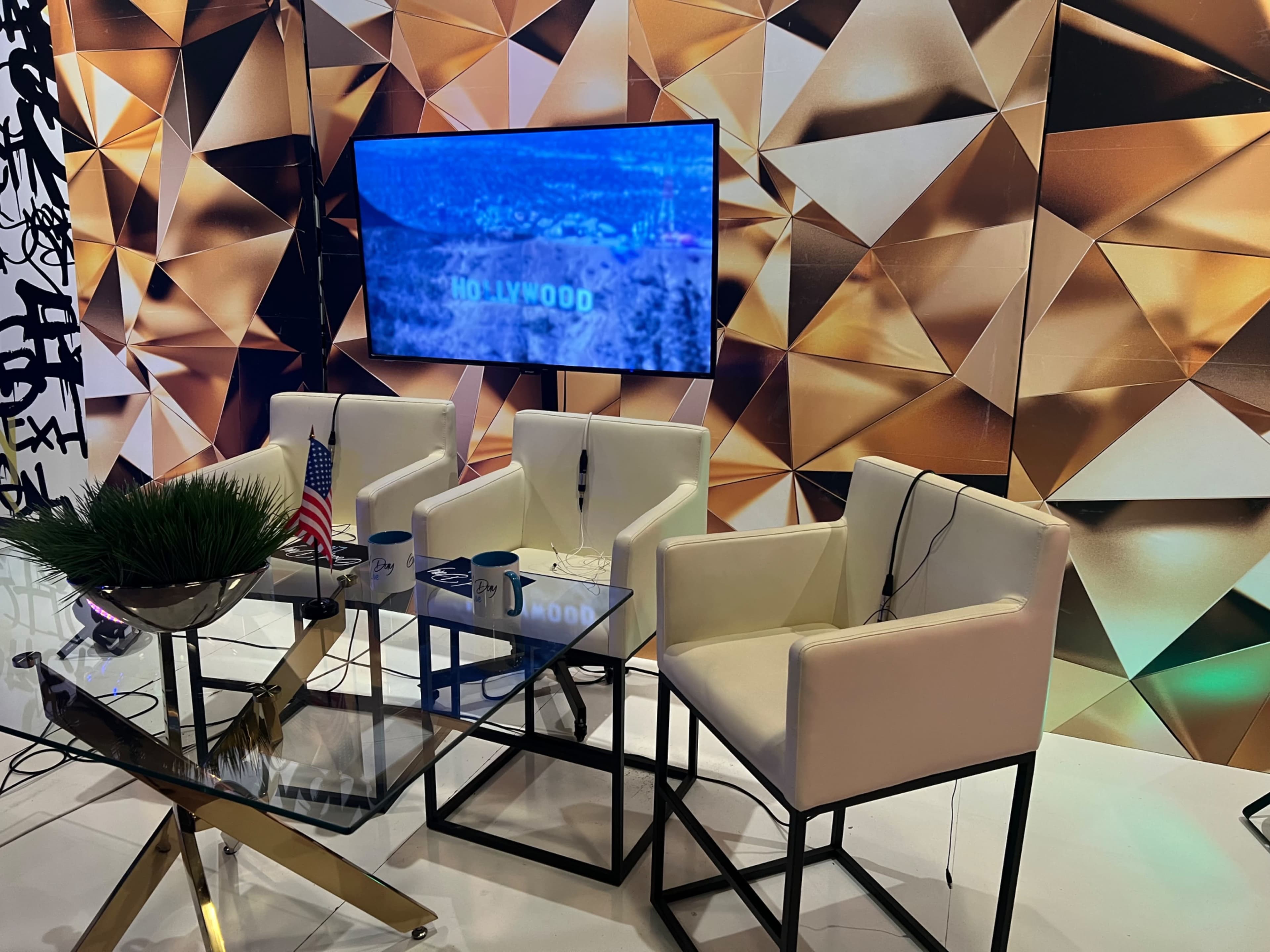 A modern interview setup featuring three white chairs, a glass table, an American flag, and a large screen displaying "HOLLYWOOD" against a geometric gold backdrop.