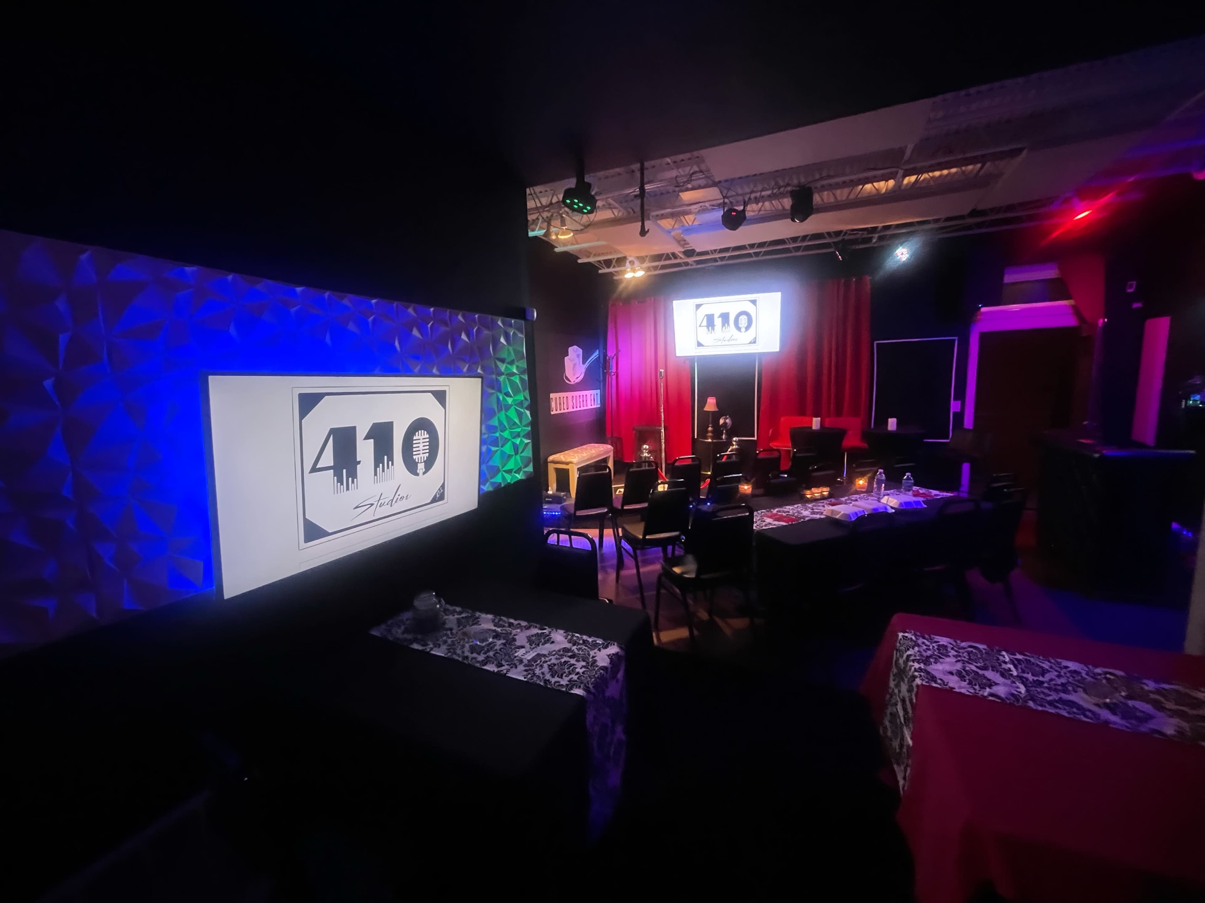 A dimly lit performance space features rows of black chairs and tables with colorful tablecloths, illuminated by various stage lights and a large banner displaying "410 Studio."