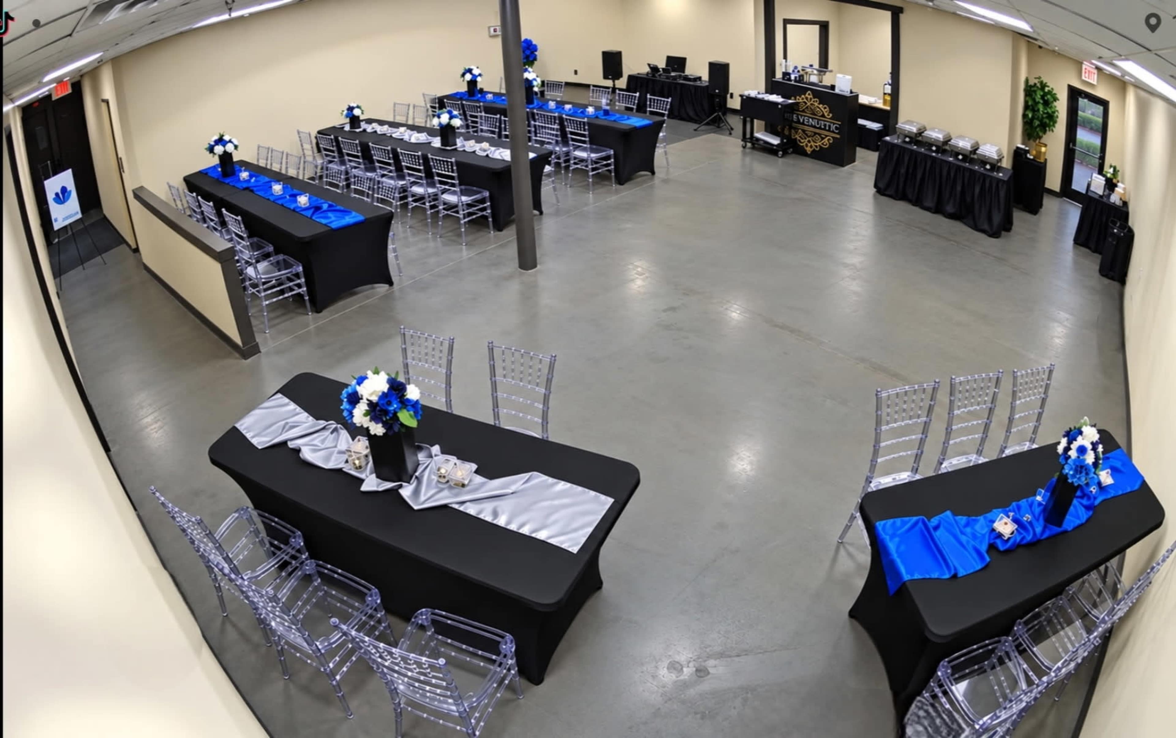 The image shows a banquet hall set up with several black and blue-themed tables arranged for an event, featuring floral centerpieces and serving stations in the background.