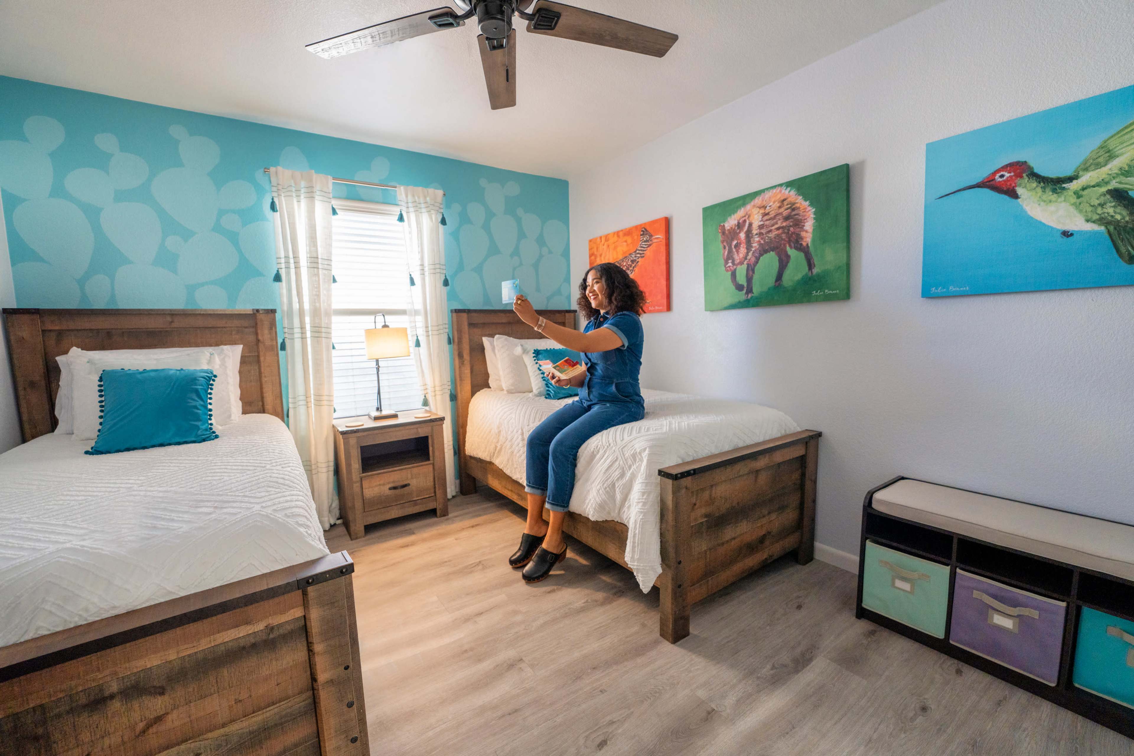 A woman sits on the edge of a wooden bed in a brightly decorated bedroom with two twin beds and colorful animal artwork on the walls.