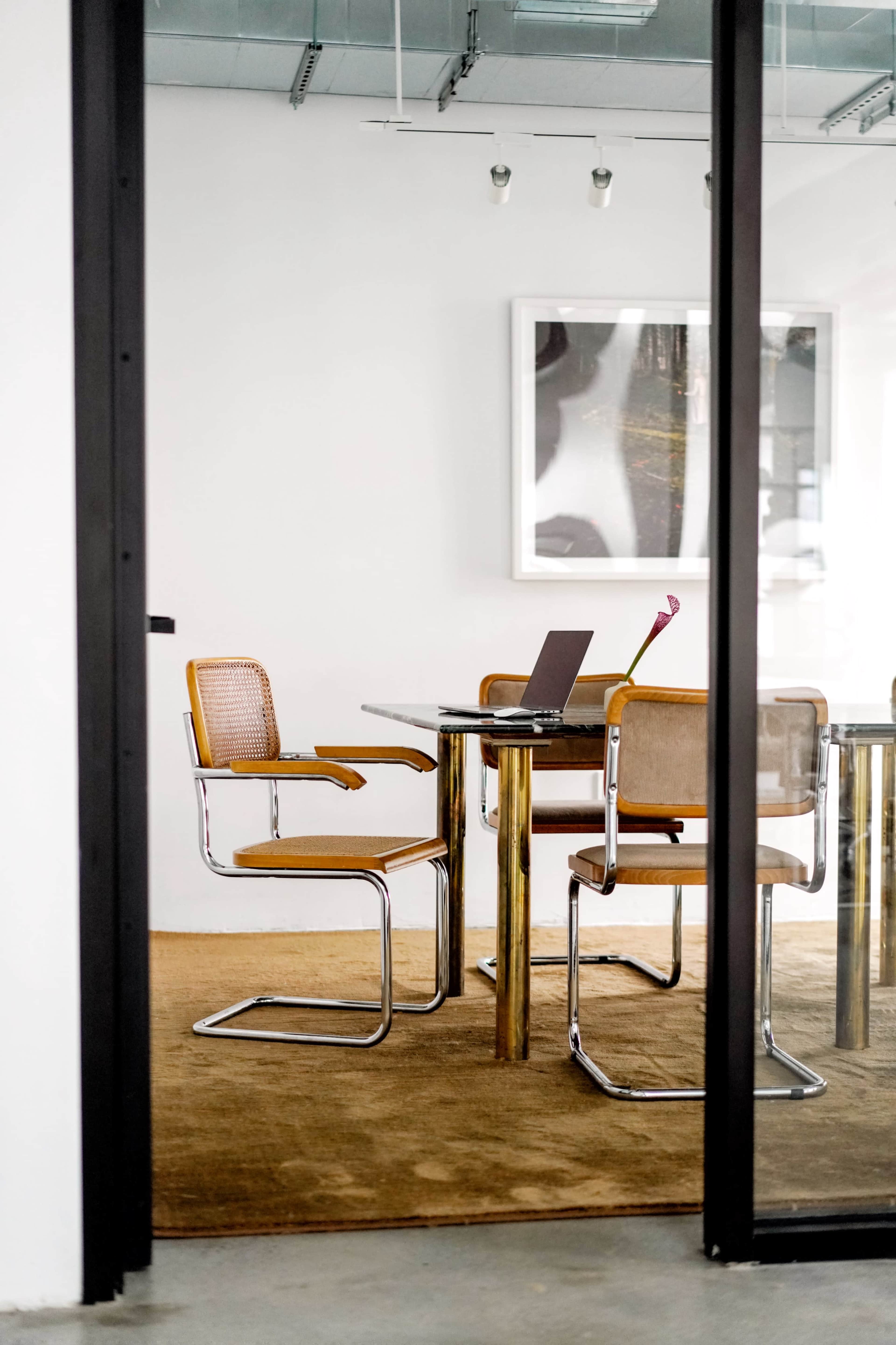 A modern office space features a glass table with a laptop and two stylish chairs on a beige carpet.