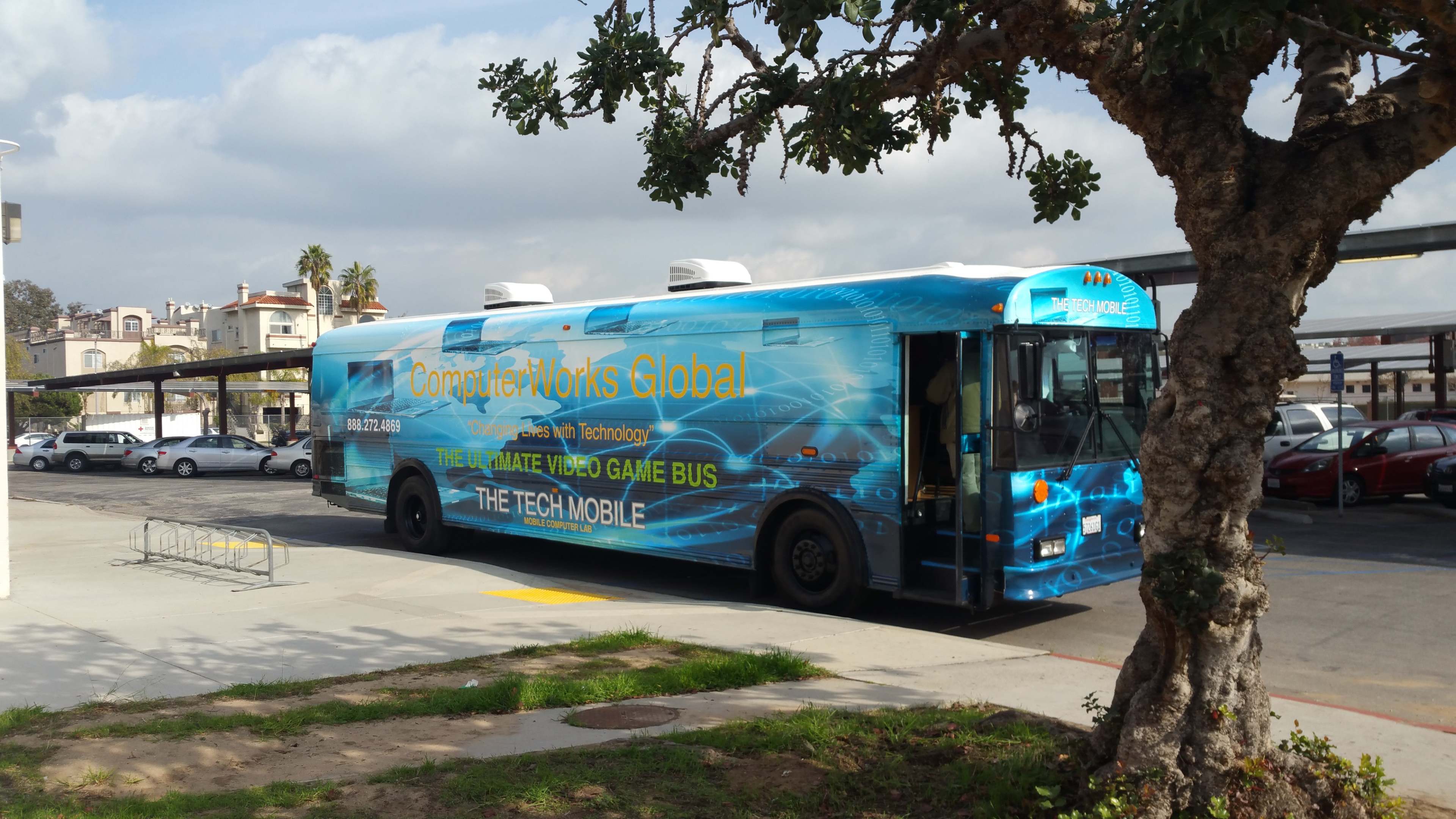 A blue bus with the text "Computer Works Global" and graphics depicting technology is parked in a lot near trees and cars.