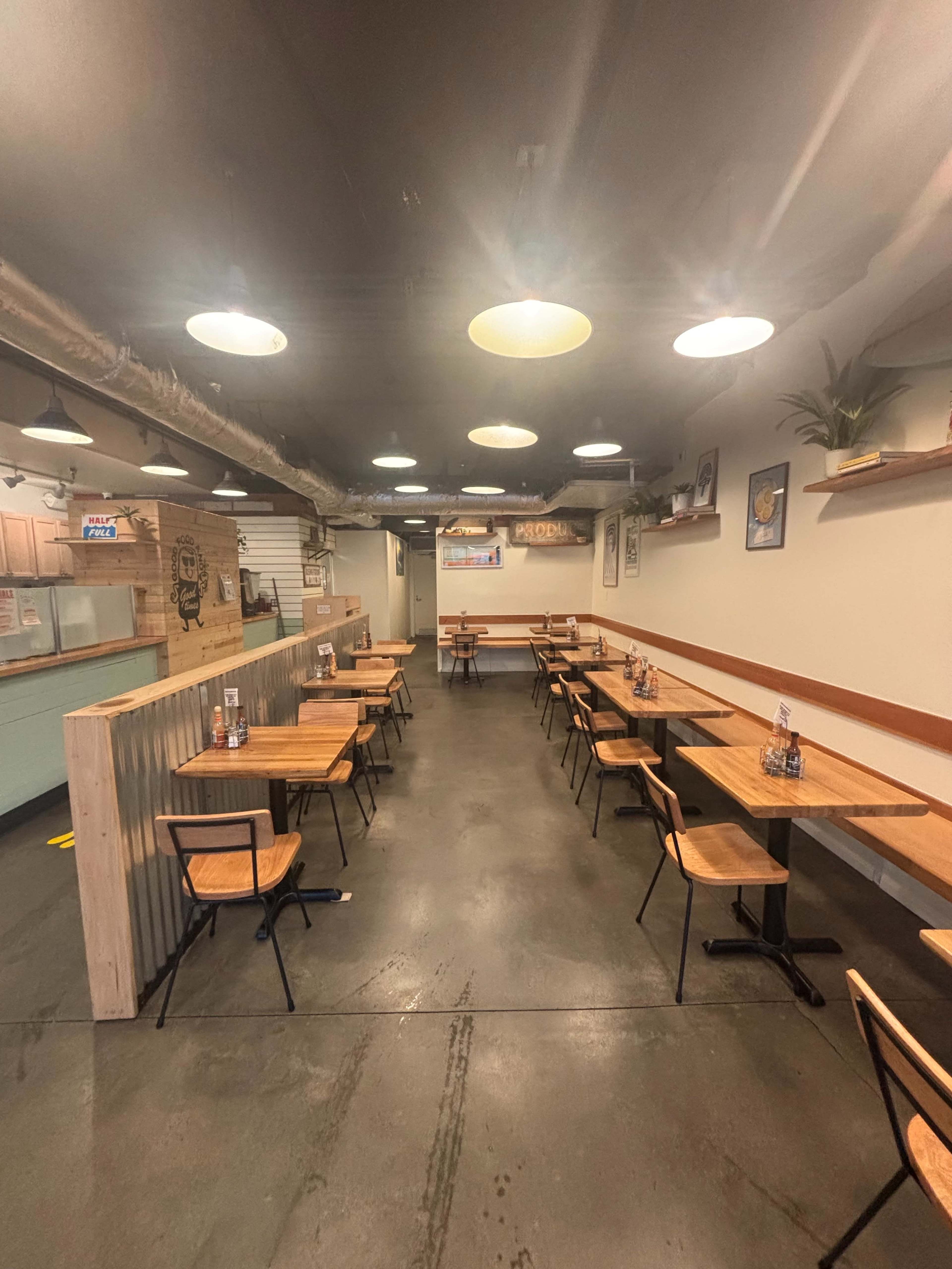 The image shows a spacious, minimalist restaurant interior featuring wooden tables and chairs arranged in rows, with soft lighting from overhead lamps.