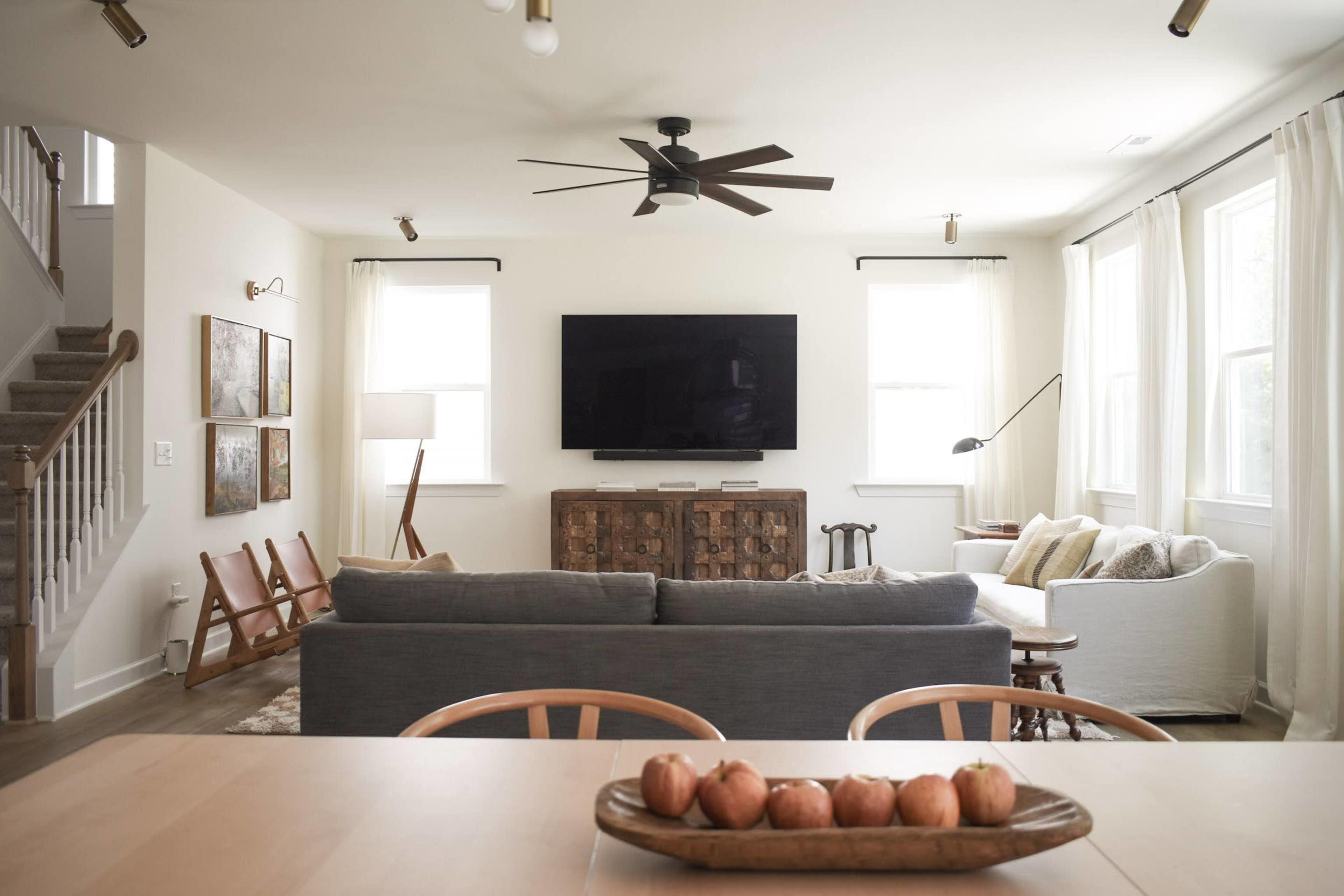 A spacious living room features a gray couch, a white sofa, a wooden coffee table with apples, and a large television mounted on the wall.