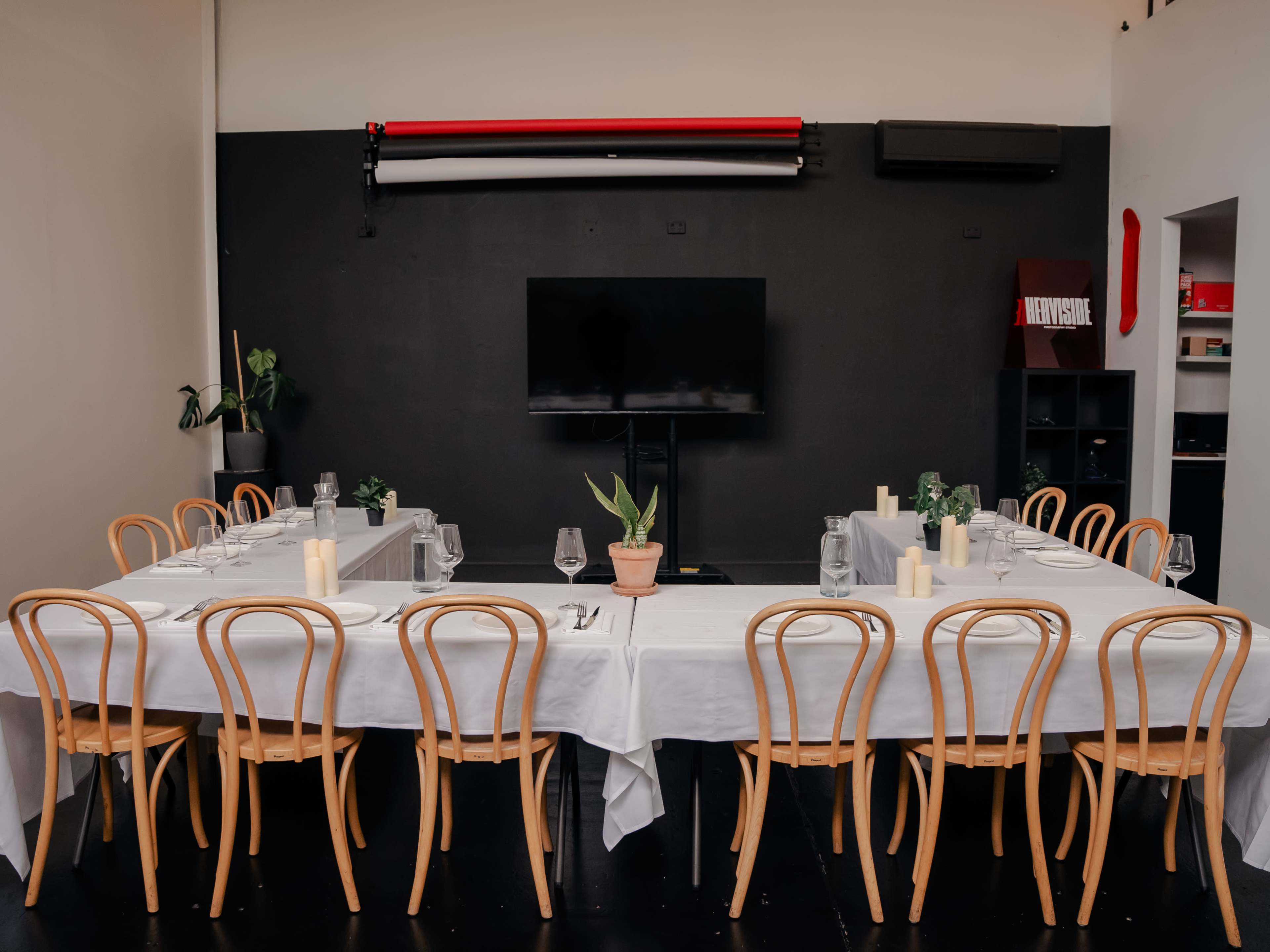 A long dining table is set with white tablecloths and elegant glassware in a modern room featuring a black wall and a large television.