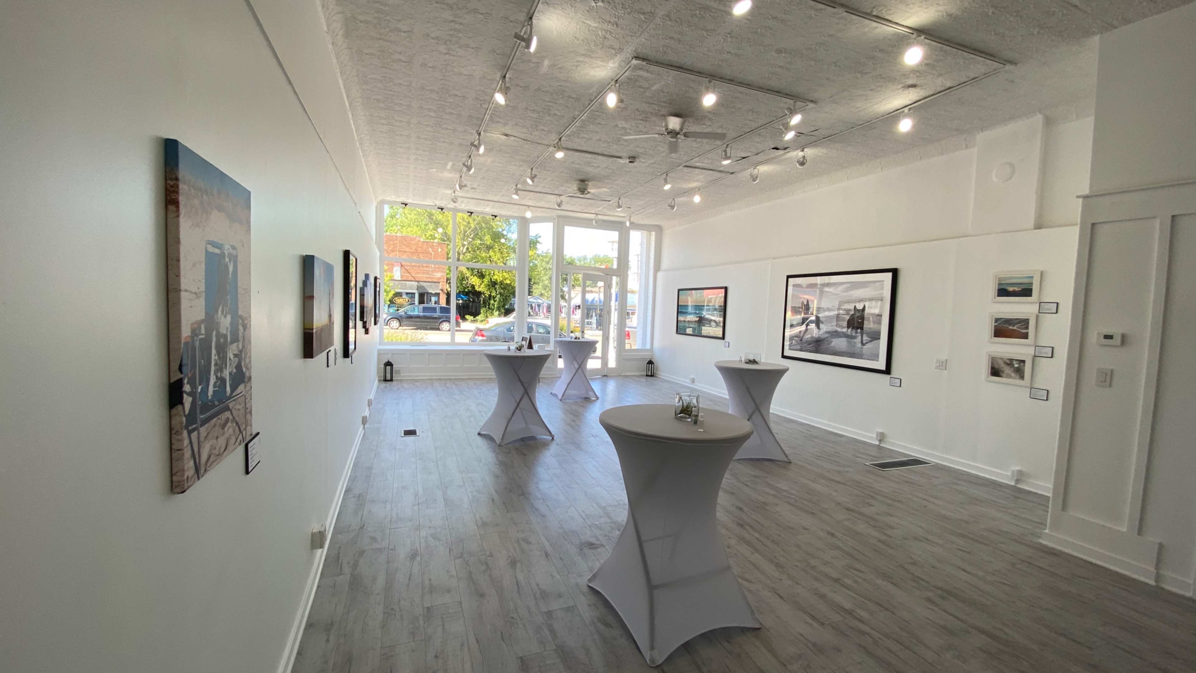 The image shows an art gallery with several framed artworks displayed on white walls, accompanied by tall tables for guests.