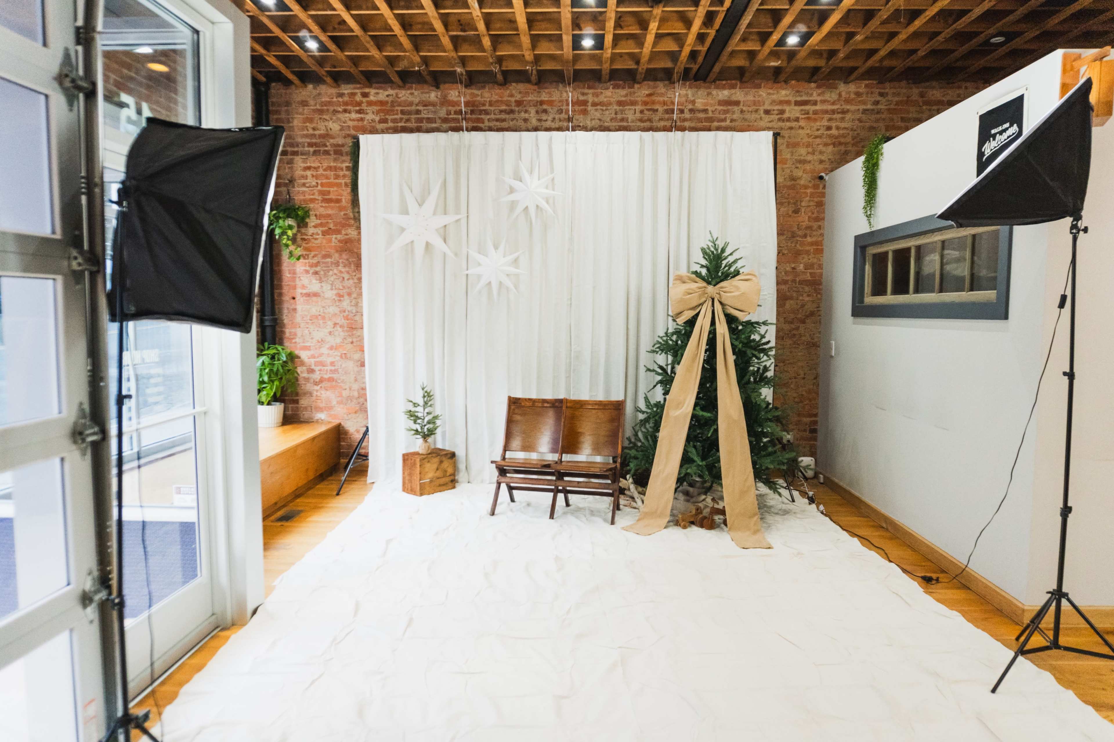 The image shows a decorated room featuring a Christmas tree with a bow, a wooden chair, and a backdrop of white fabric and paper stars.