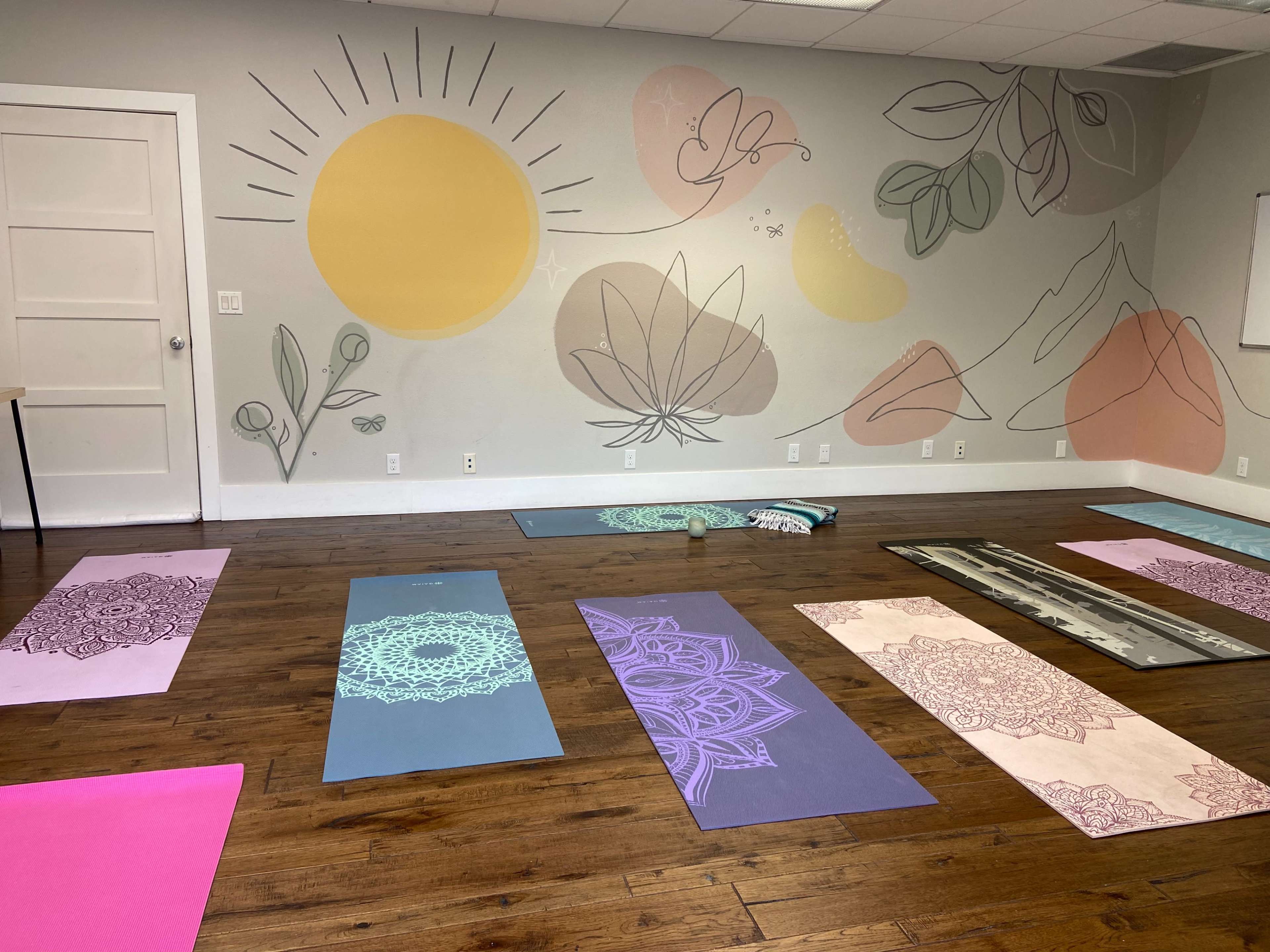 A yoga studio features several mats arranged on a wooden floor with a mural of sun and plant motifs on the wall.