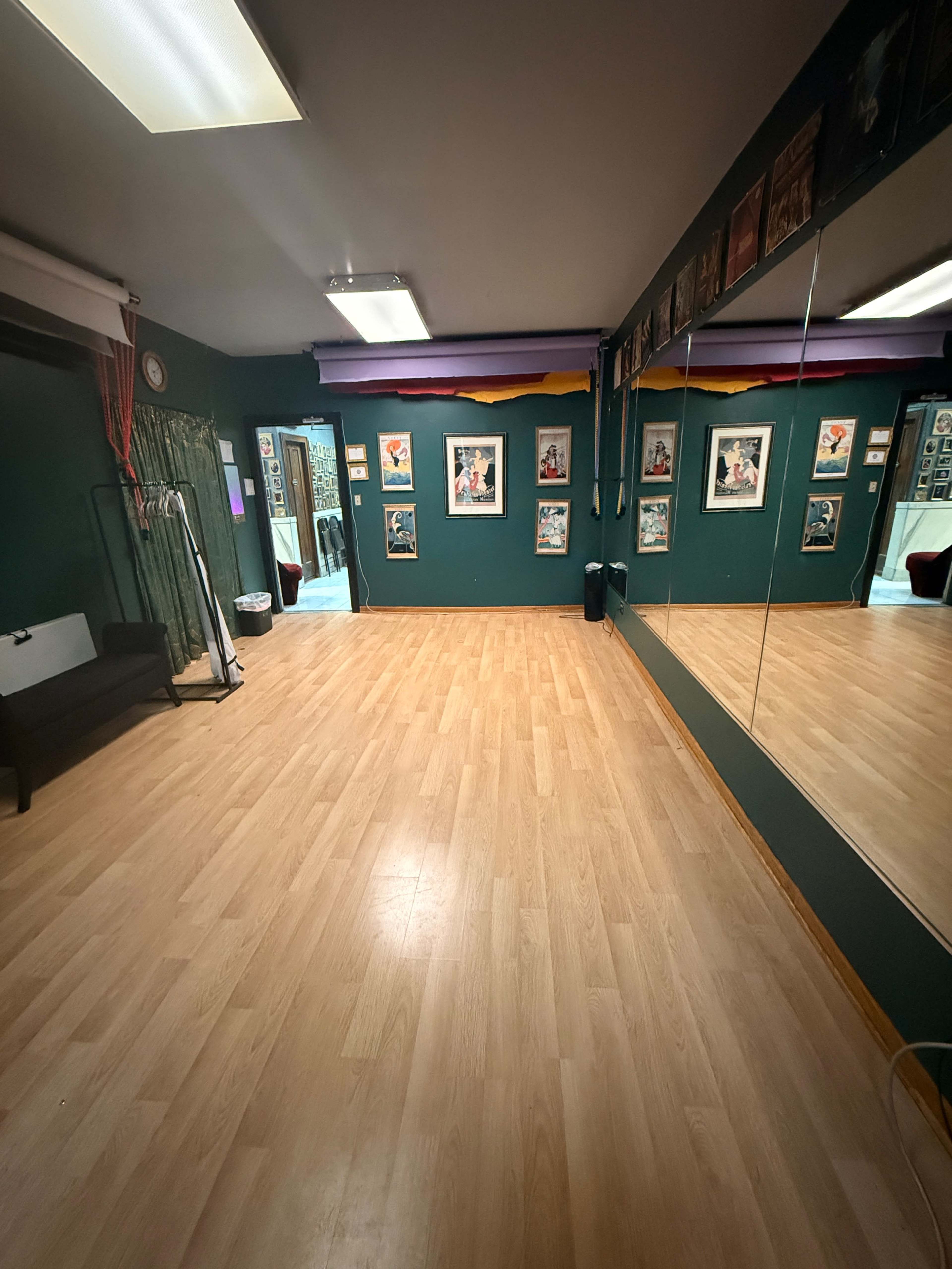 The image shows a spacious and well-lit dance studio with wooden flooring, mirrored walls, and framed artworks displayed on the green walls.