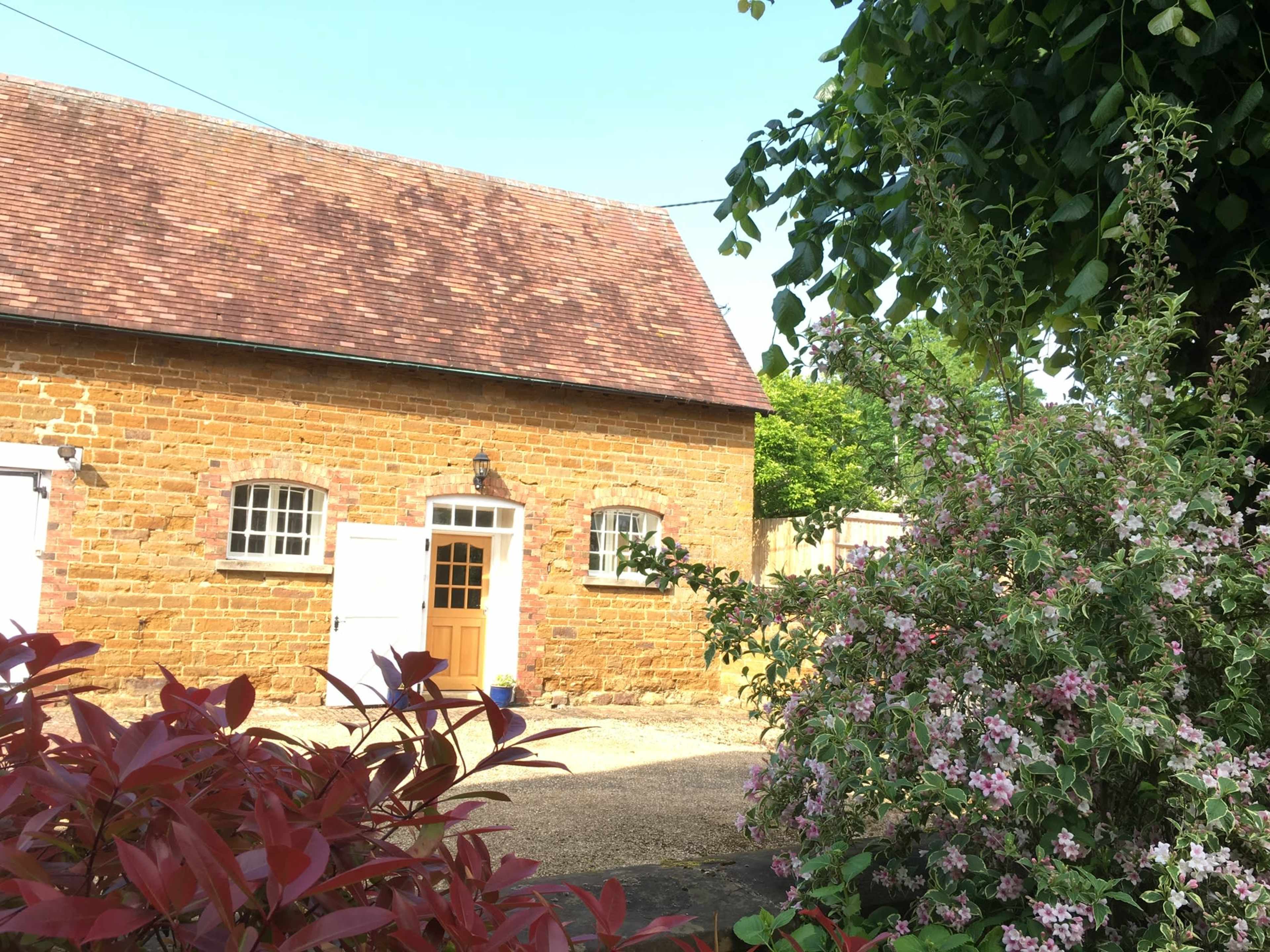 A stone cottage with a brown roof is surrounded by flowering plants and greenery.