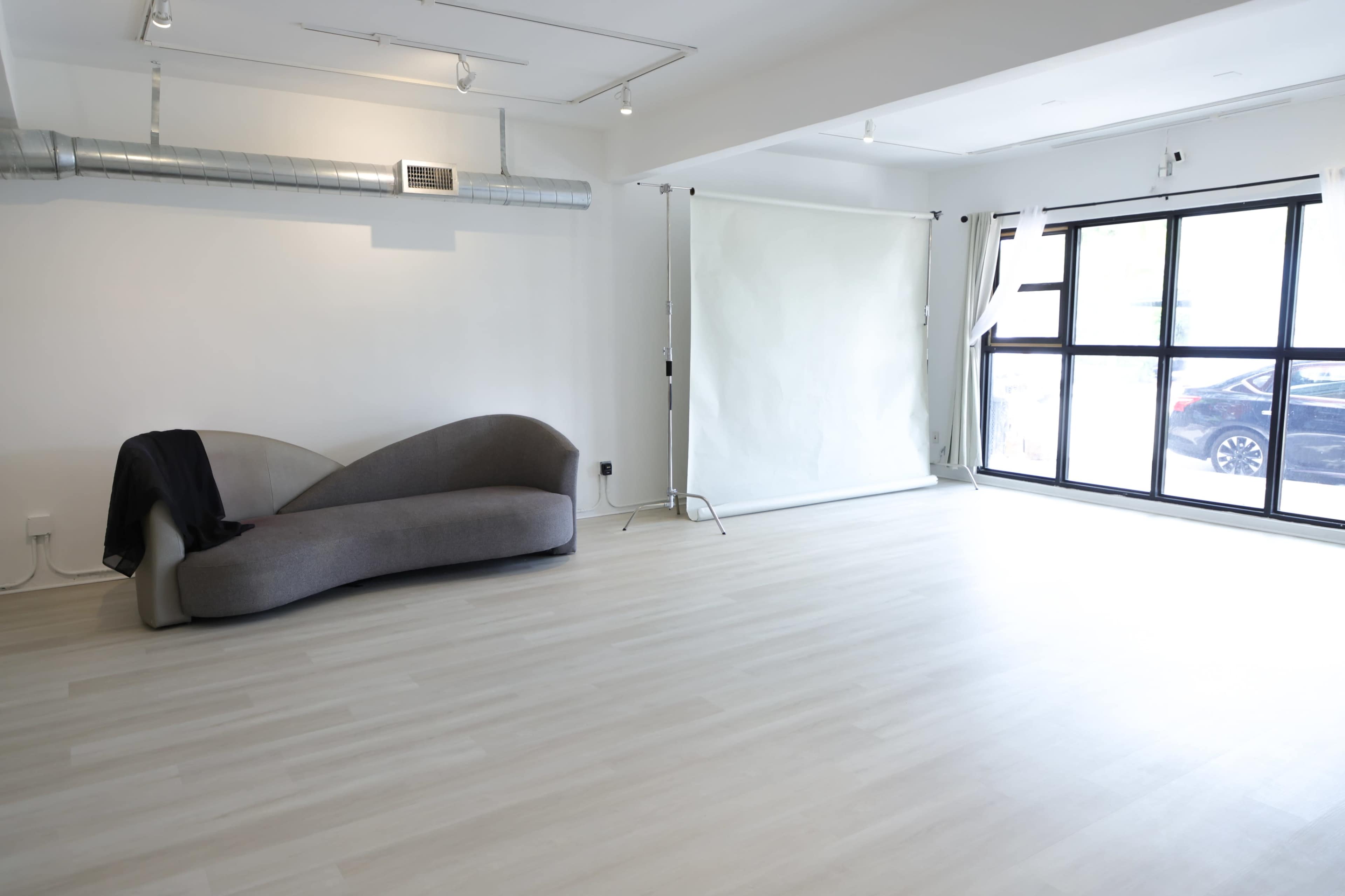 The image shows a spacious, well-lit room featuring a gray couch in one corner and a large backdrop setup near a window.