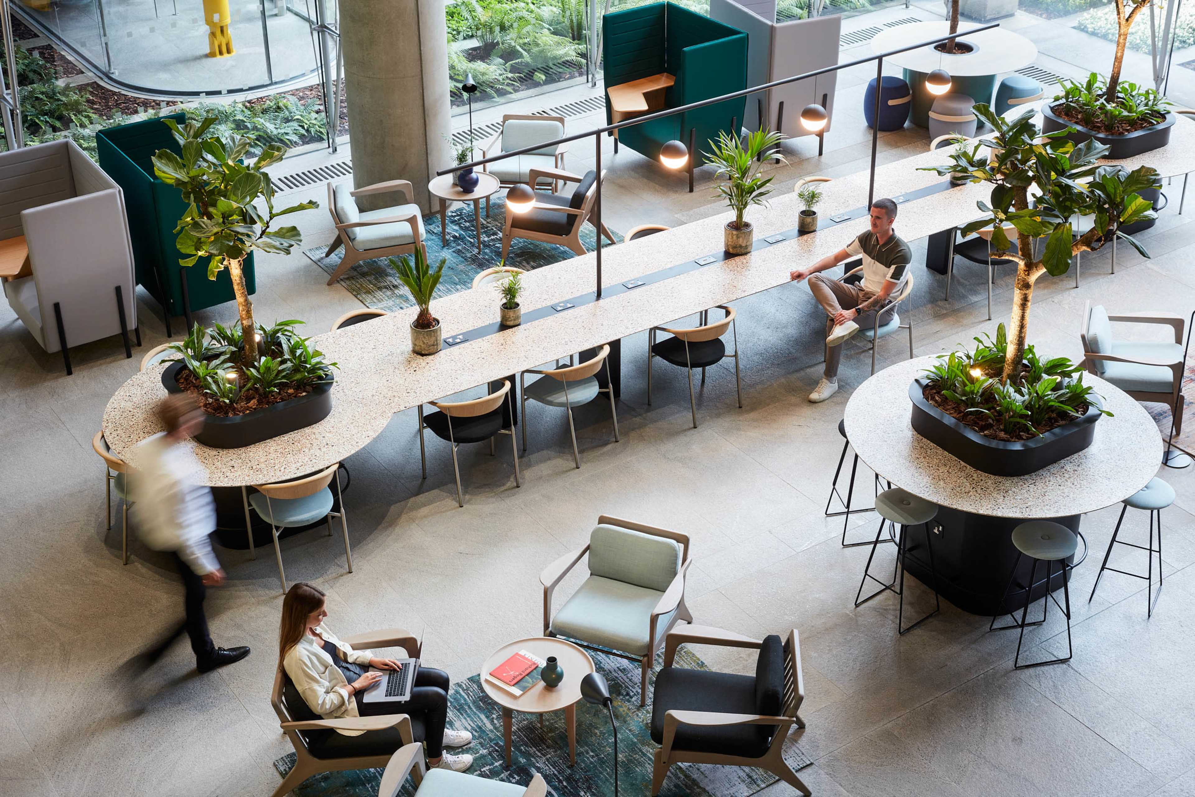 The image shows a modern, open workspace featuring long tables with potted plants, surrounded by chairs, where two people are engaged in different activities.