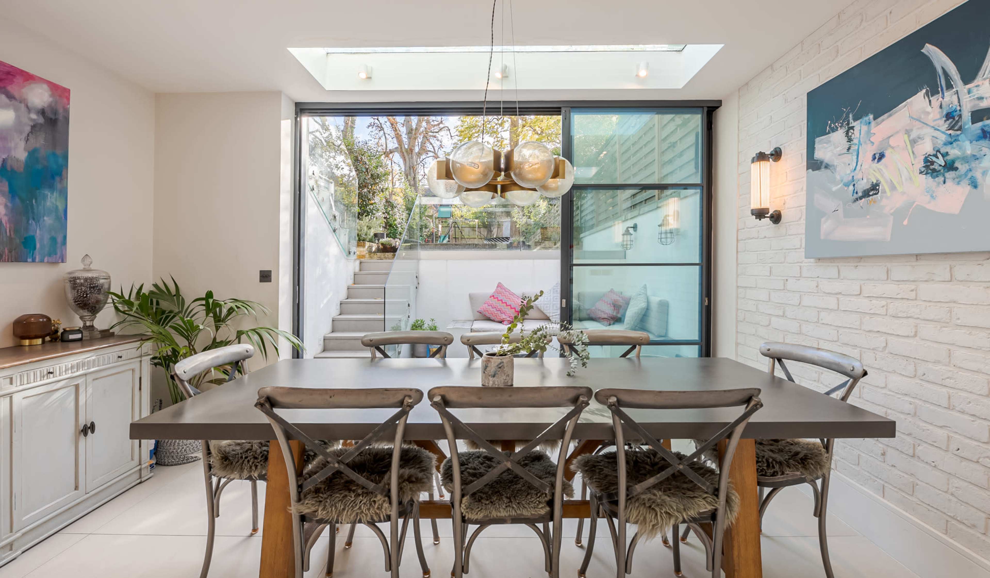 A dining area features a long table surrounded by wooden chairs, with a large glass wall revealing a staircase and outdoor space in the background.