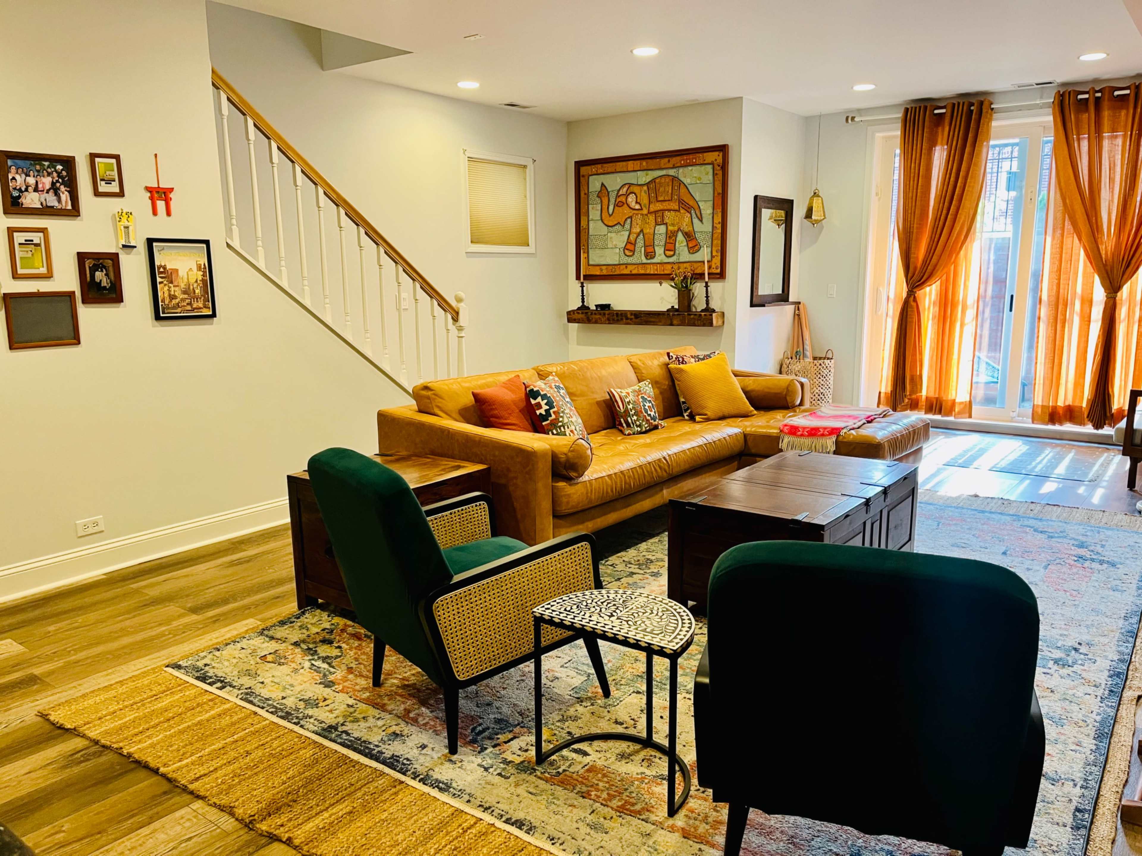 The living room features a tan sofa with colorful cushions, two green chairs, a wooden coffee table, and large windows dressed in orange curtains.