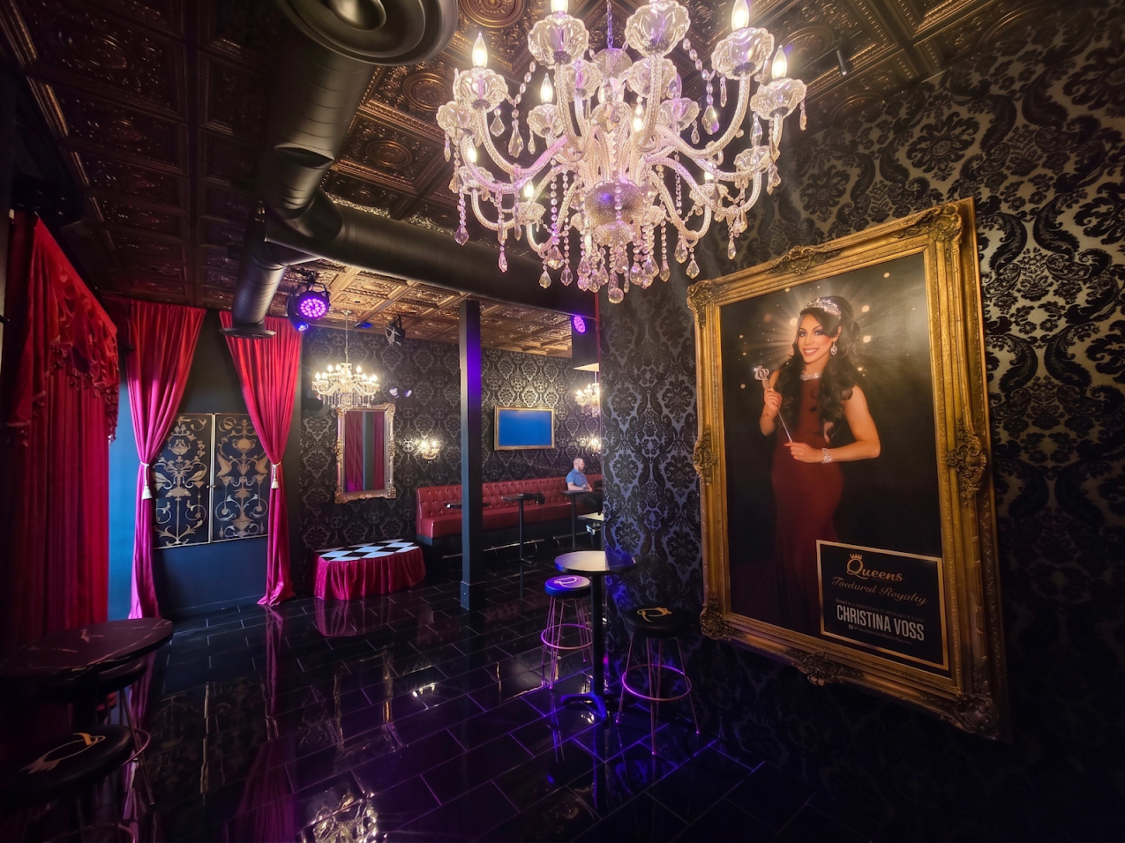The image shows an elegantly decorated interior of a venue featuring a chandelier, ornate wallpaper, and a framed portrait on the wall.