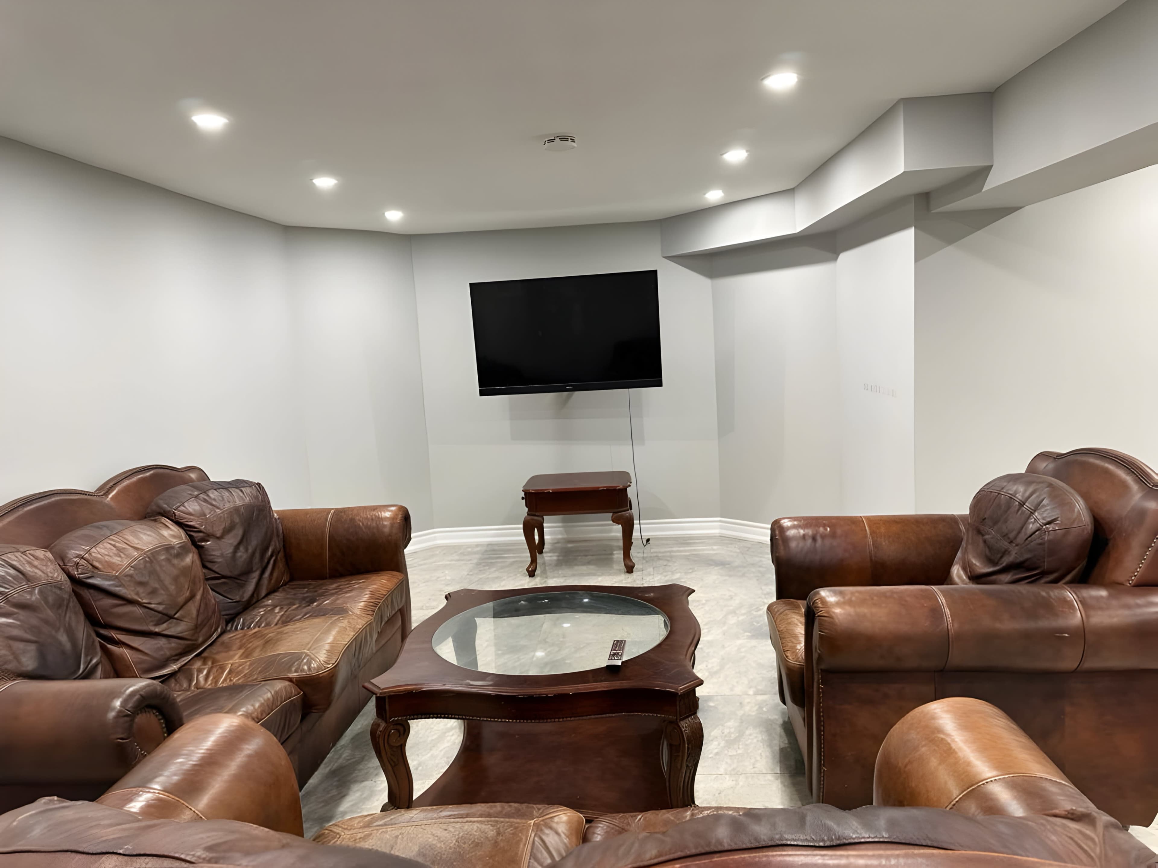A basement living area with brown leather sofas arranged around a coffee table and a wall-mounted television.