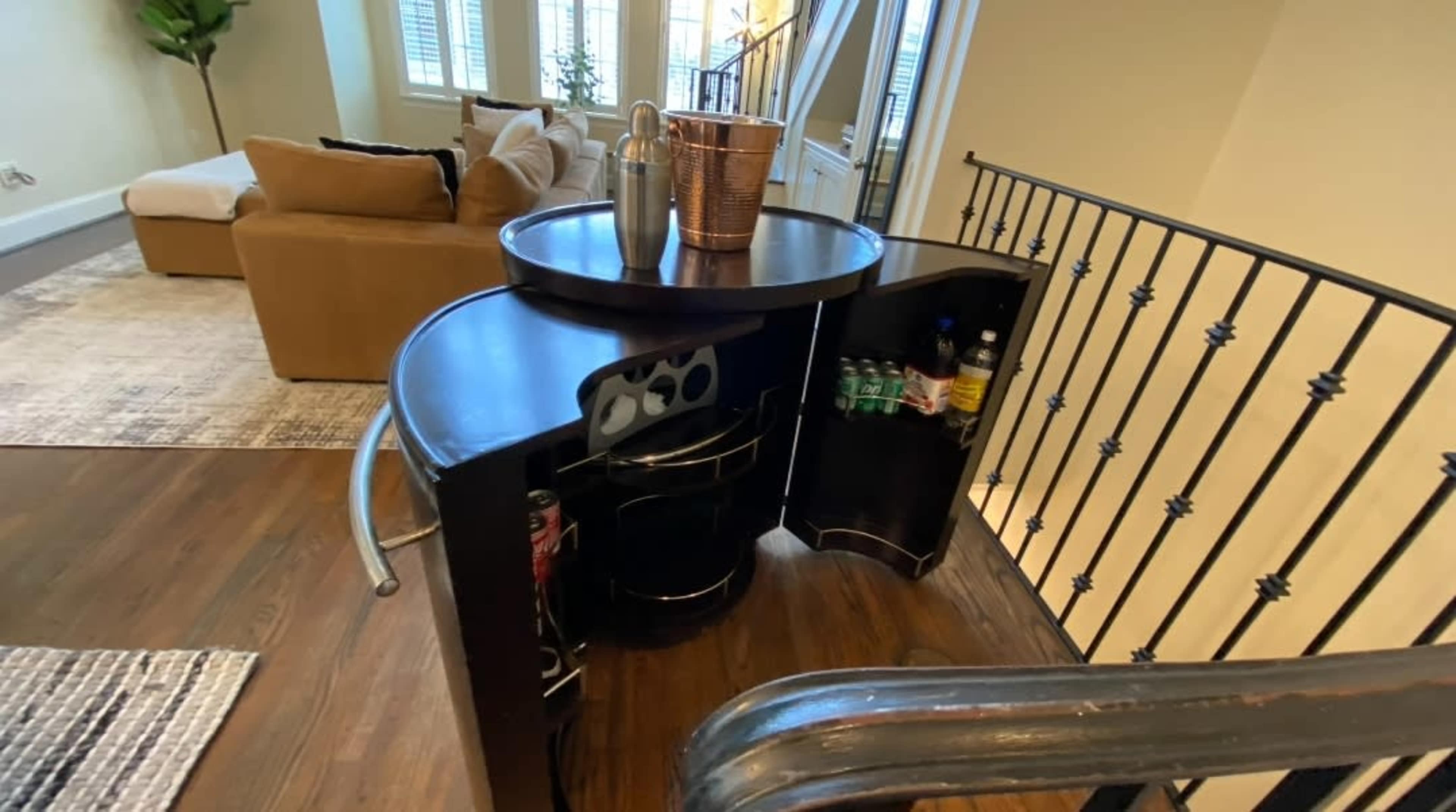 A home bar setup with a circular top, accompanied by a basket and several beverage bottles, is positioned next to a railing in a spacious living area.