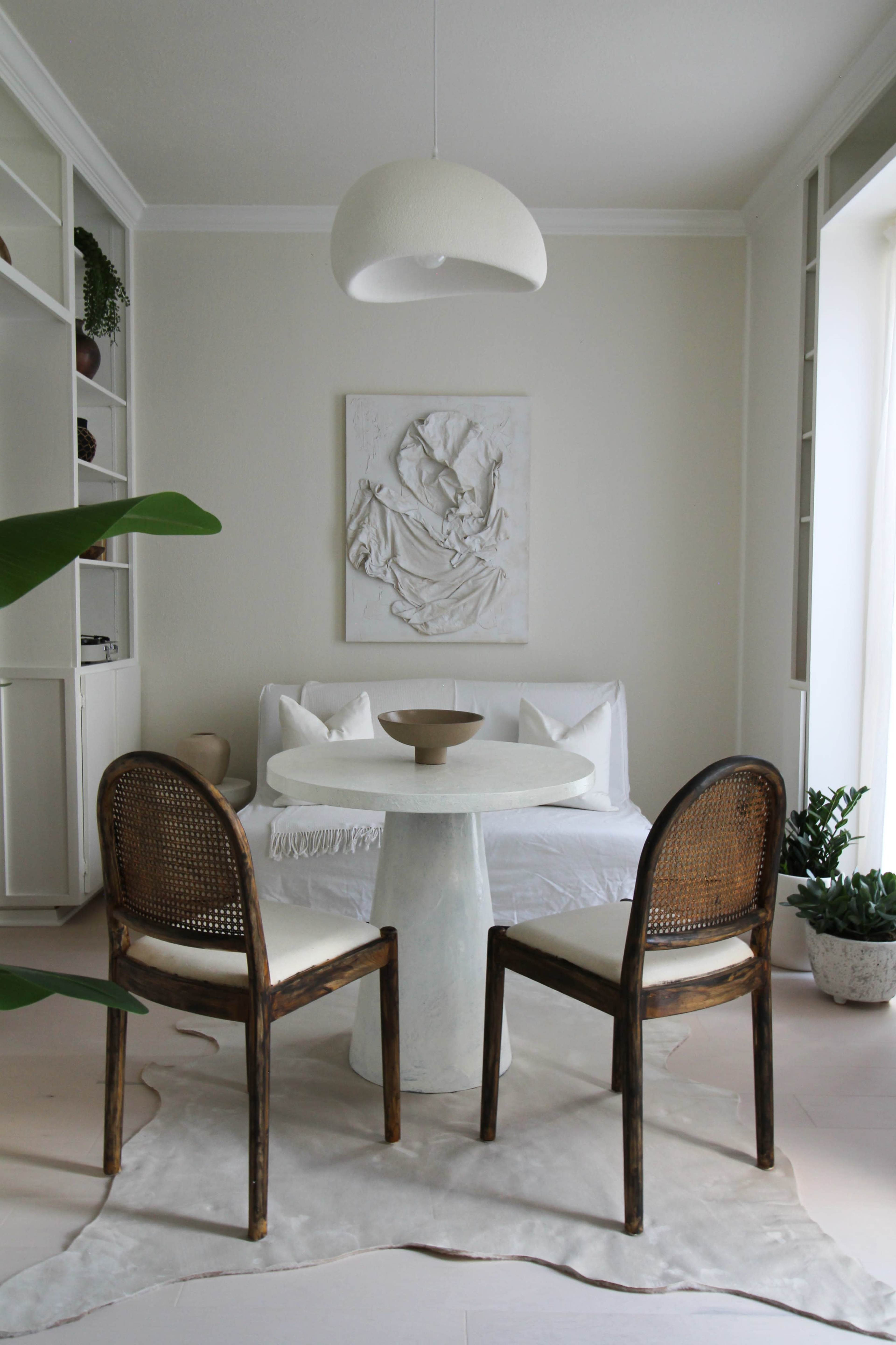 A modern dining area features a round table with a white pedestal, two woven chairs, and a minimalist wall art piece.