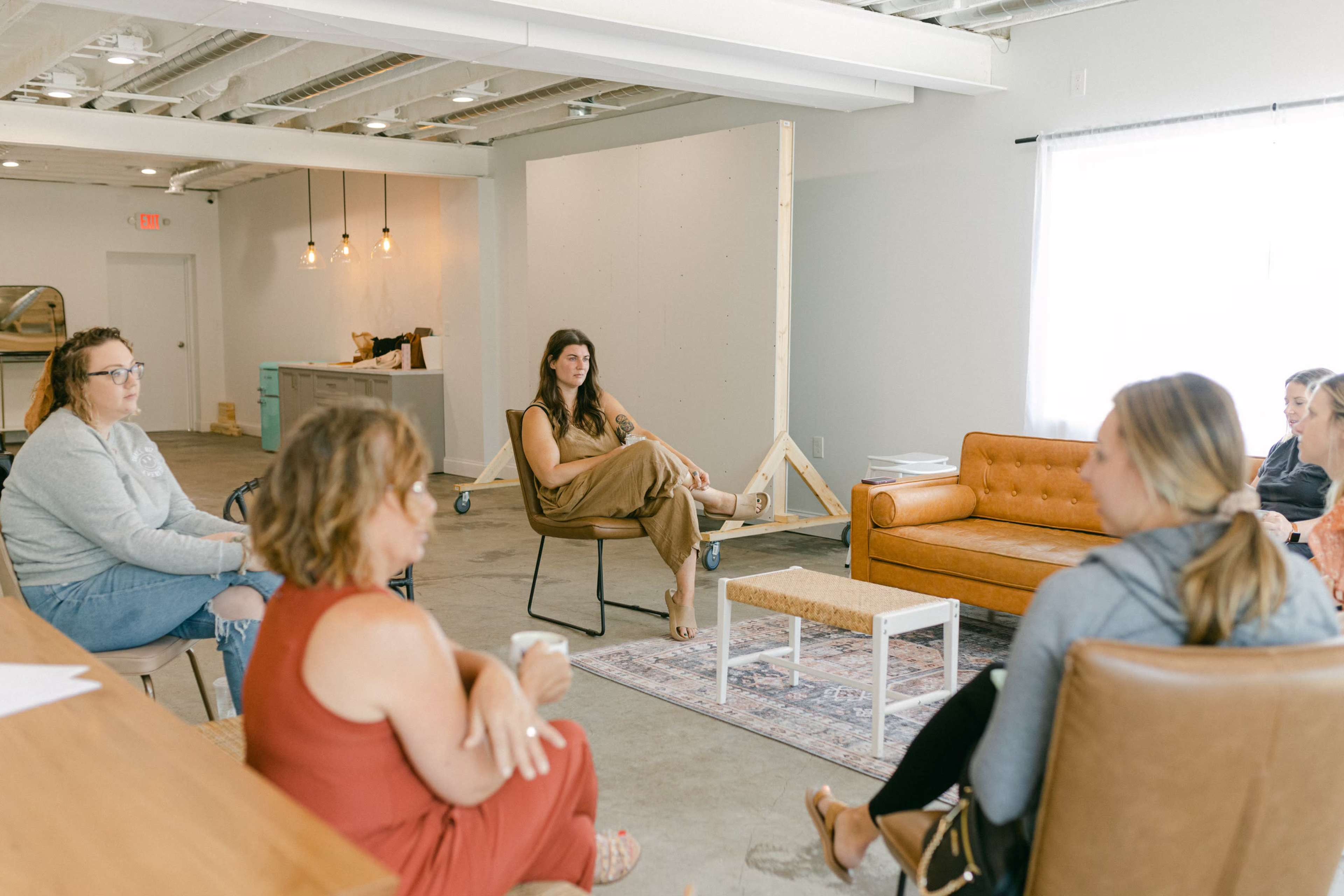 A group of six people are seated in a light-filled room, engaging in conversation, with a large wooden board in the background and a few couches arranged around a small rug.