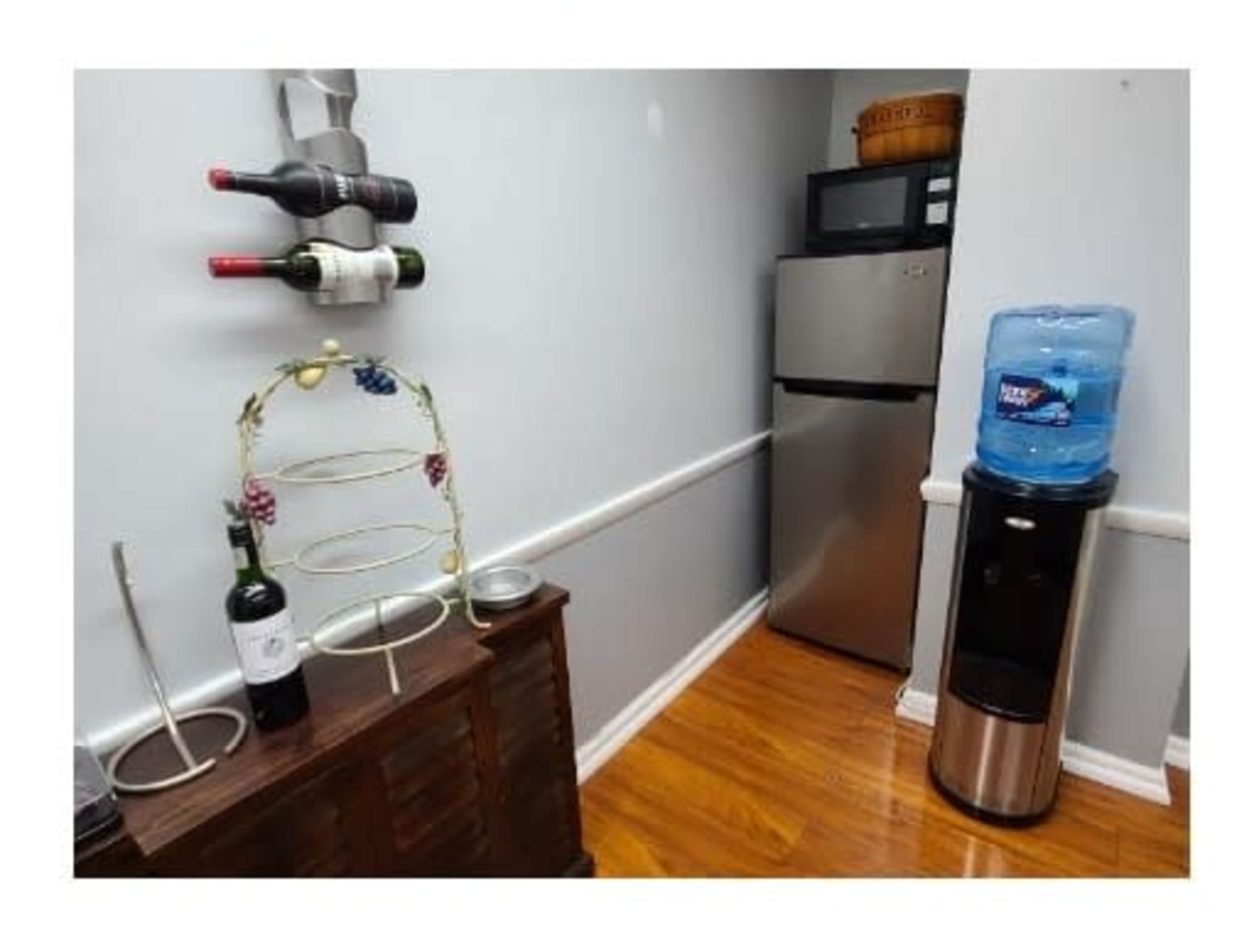The image shows a kitchen corner with a wine rack holding three bottles, a stainless steel refrigerator, and a water cooler next to a wooden cabinet.